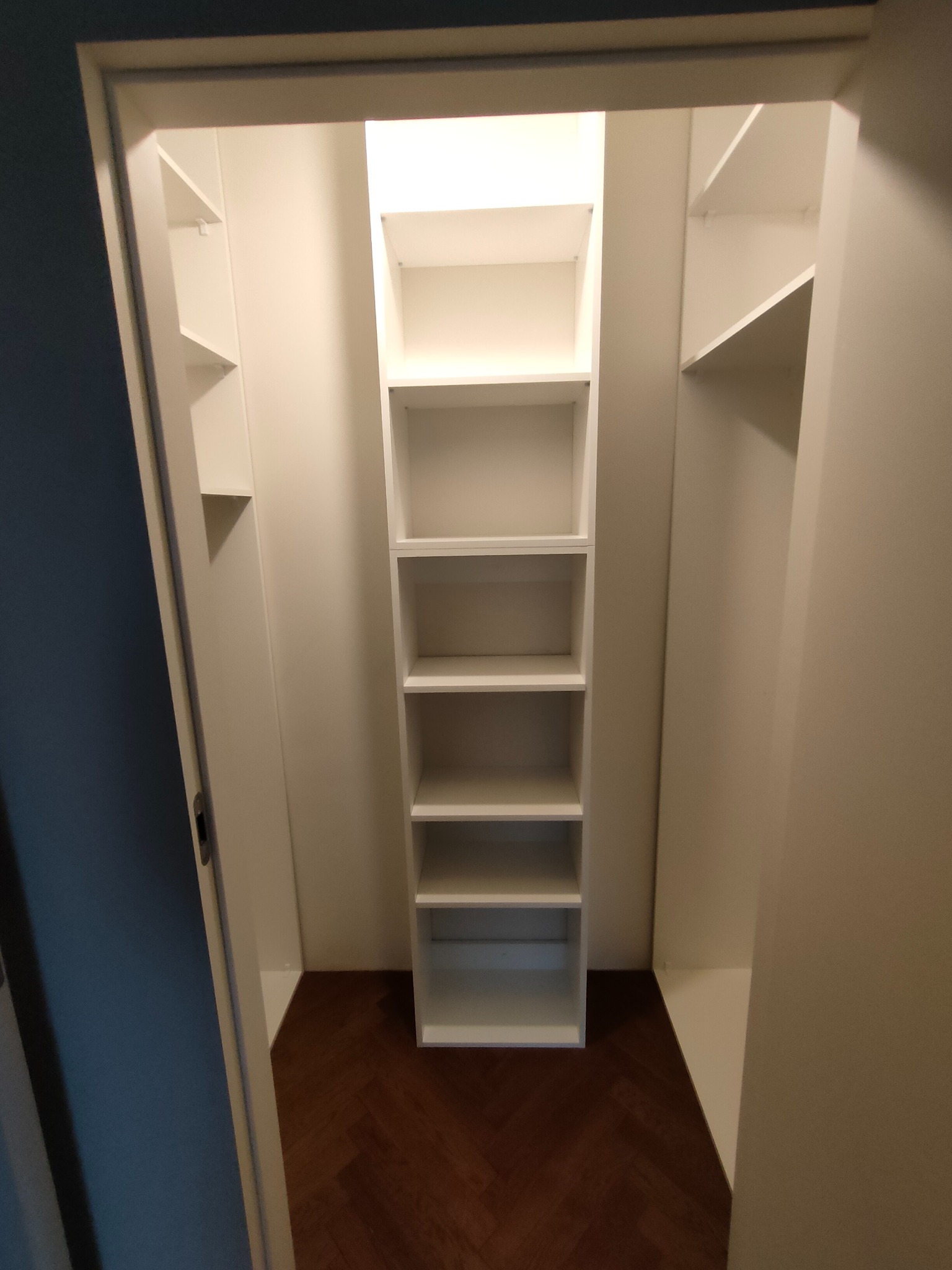 Wnętrze garderoby z białymi półkami na wymiar i parkietem w jodełkę. Widok z korytarza, drzwi uchylone. Minimalistyczny design, jasne oświetlenie.