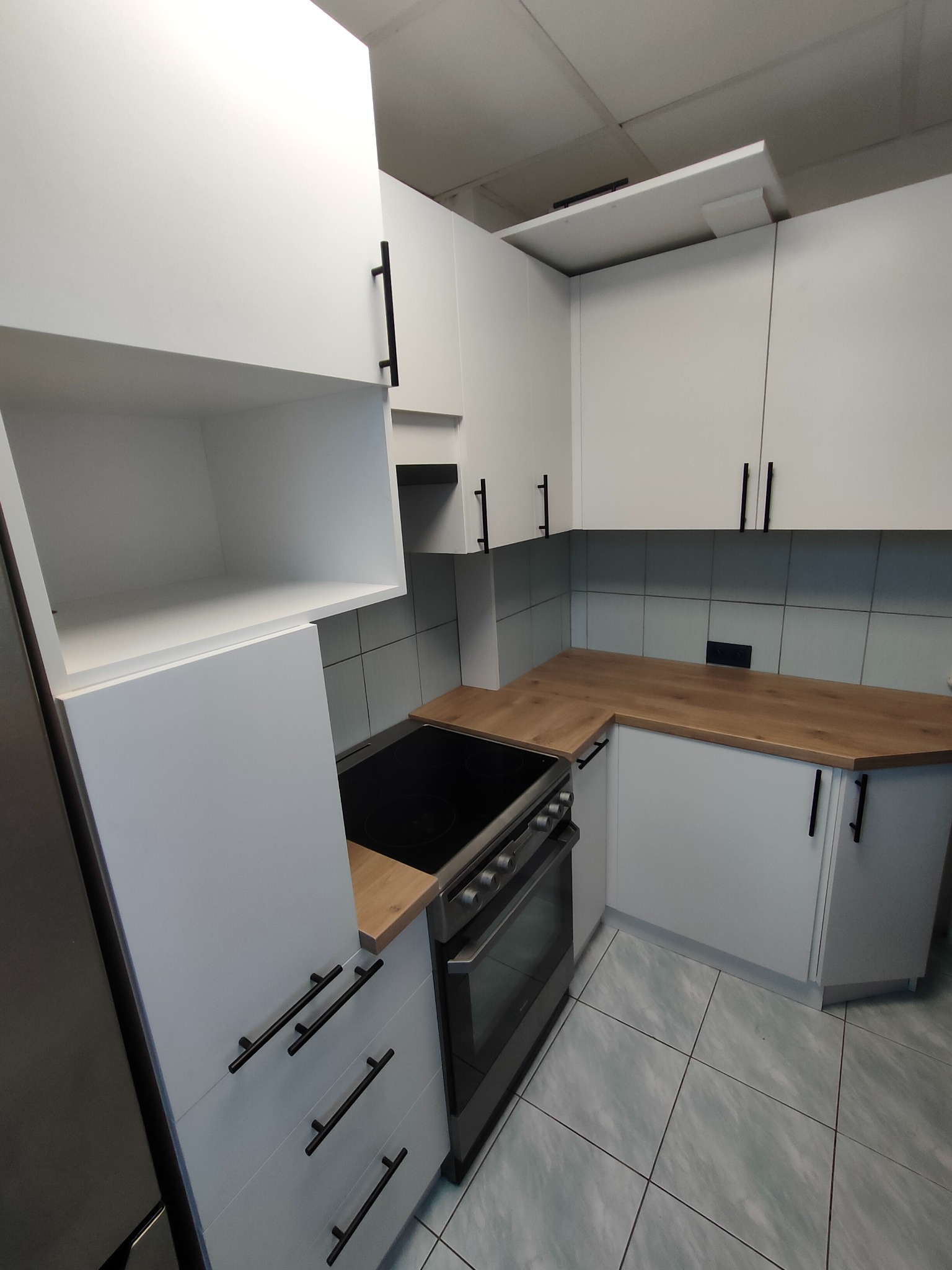 Nowoczesna kuchnia z białymi szafkami i drewnianym blatem. Czarne, podłużne uchwyty dodają elegancji. Widoczna płyta grzewcza i piekarnik. Jasne płytki na podłodze i ścianie.