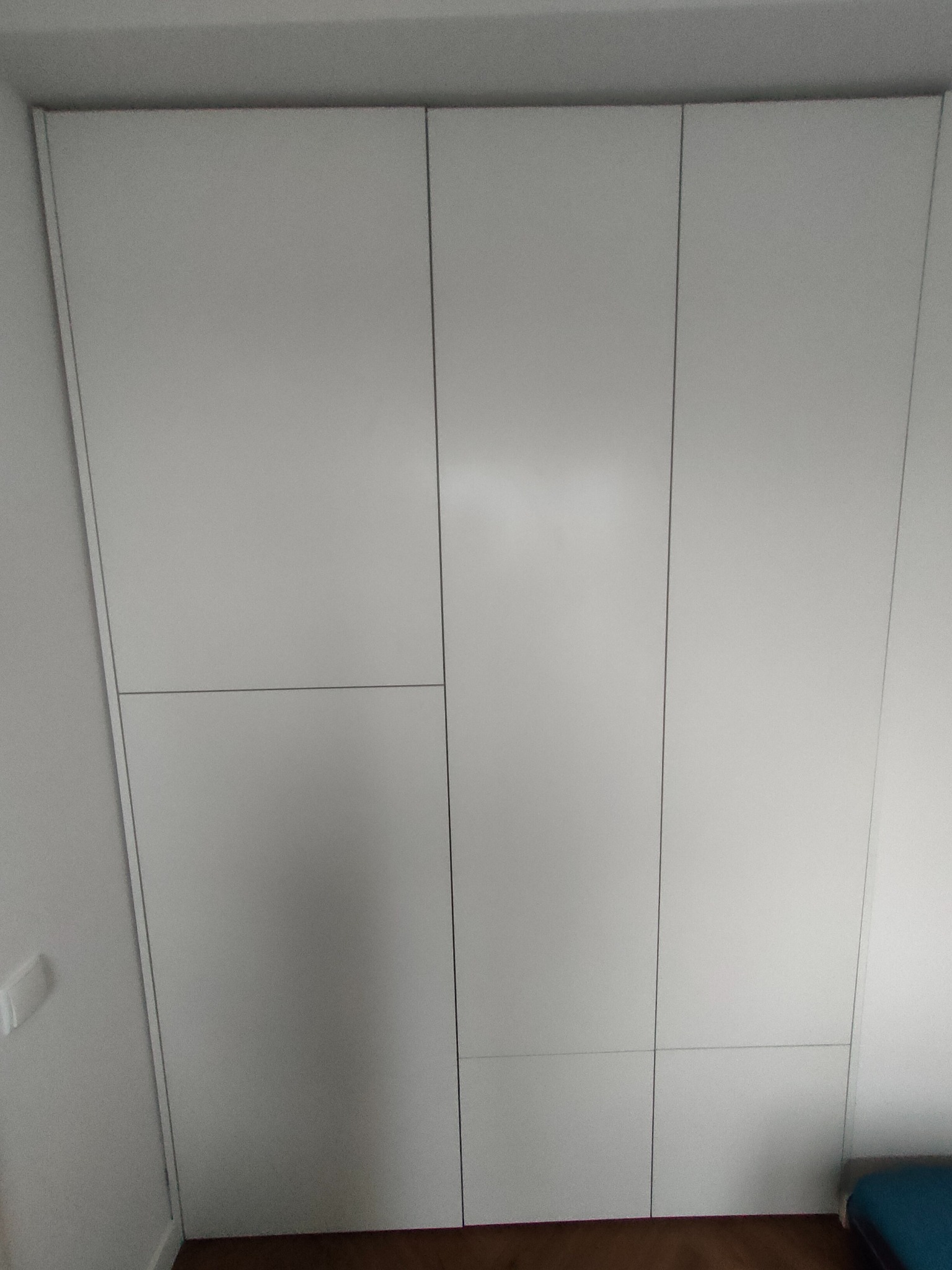 Biała szafa wnękowa z minimalistycznymi drzwiami, widoczna z dołu, z delikatnym oświetleniem i drewnianą podłogą. Układ paneli tworzy asymetryczny wzór.