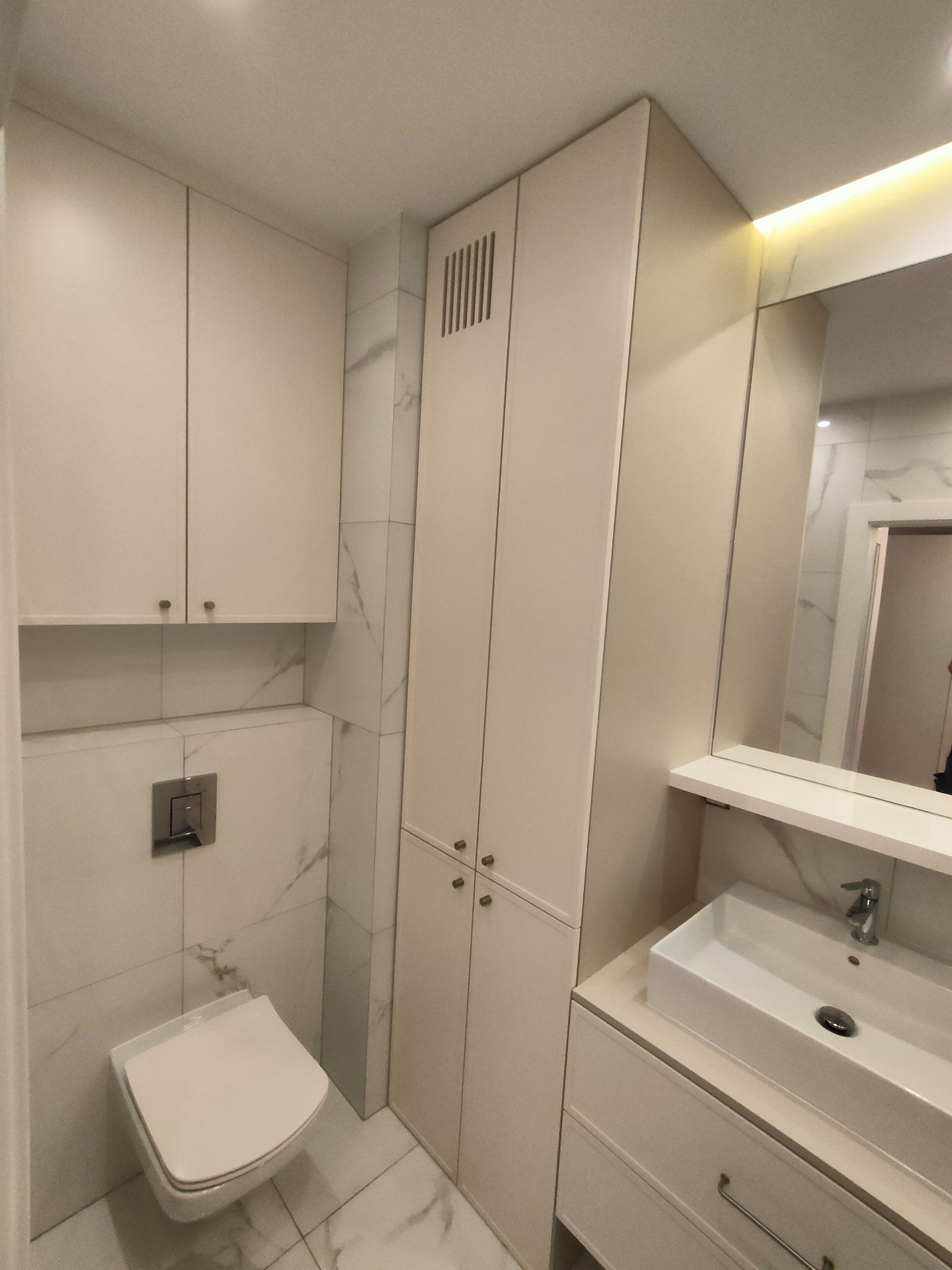 Jasna łazienka z zabudową meblową na wymiar: szafki wiszące, słupek z lamelami, umywalka z szufladami. Marmurowe płytki na ścianach, podświetlane lustro.