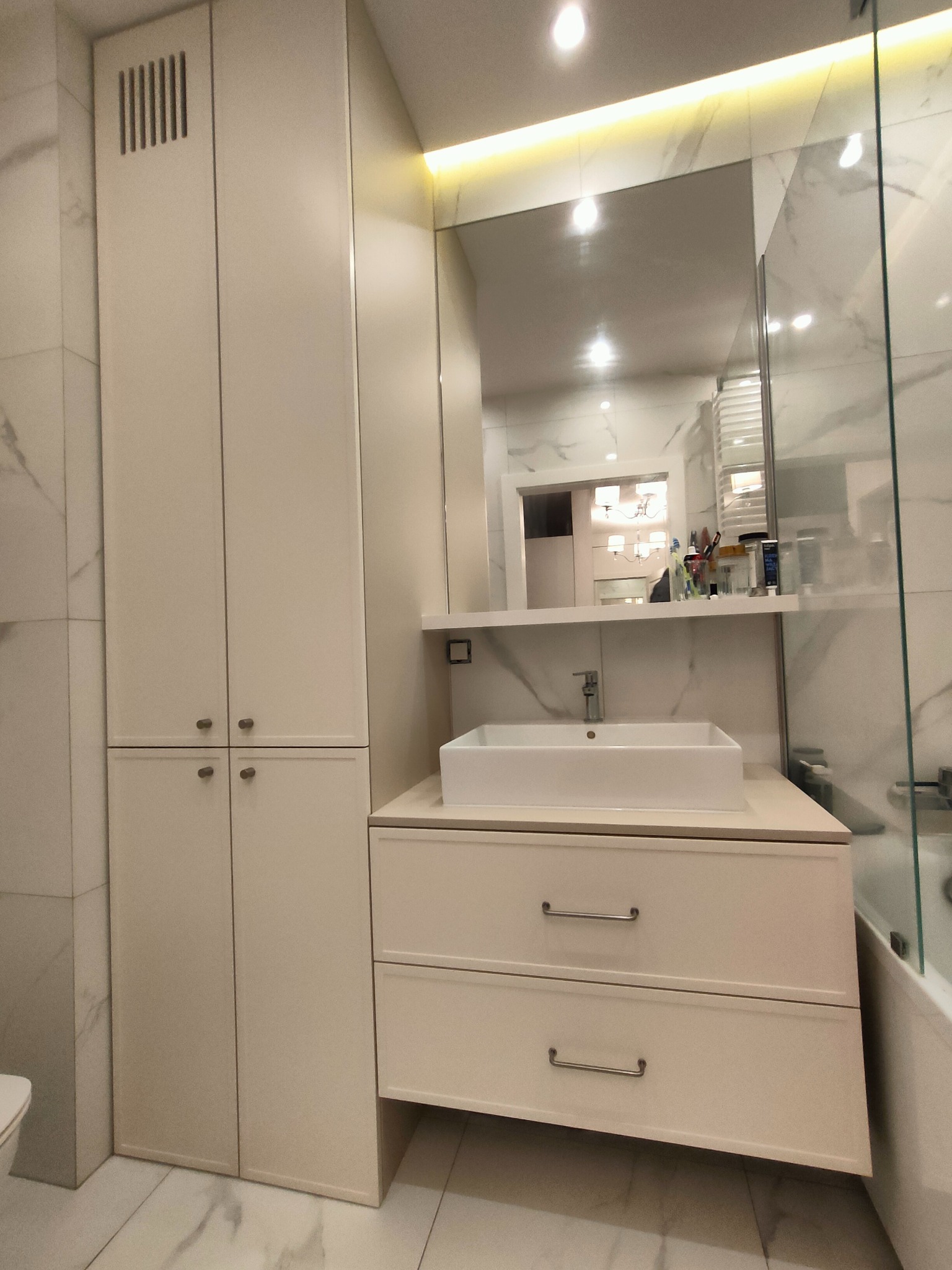 Jasna łazienka z szafką pod umywalką i wysoką szafą w zabudowie. Lustro z oświetleniem LED. Widoczne marmurowe płytki i fragment kabiny prysznicowej.