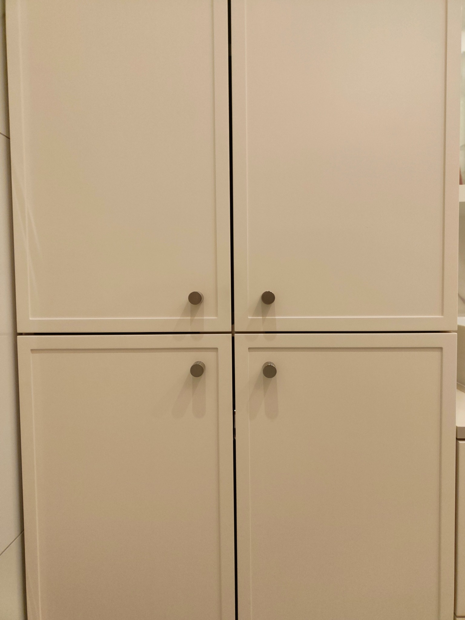 Zamknięte, kremowe fronty szafy z czterema drzwiczkami i okrągłymi, metalowymi uchwytami. Minimalistyczny design, gładkie powierzchnie, subtelne oświetlenie.