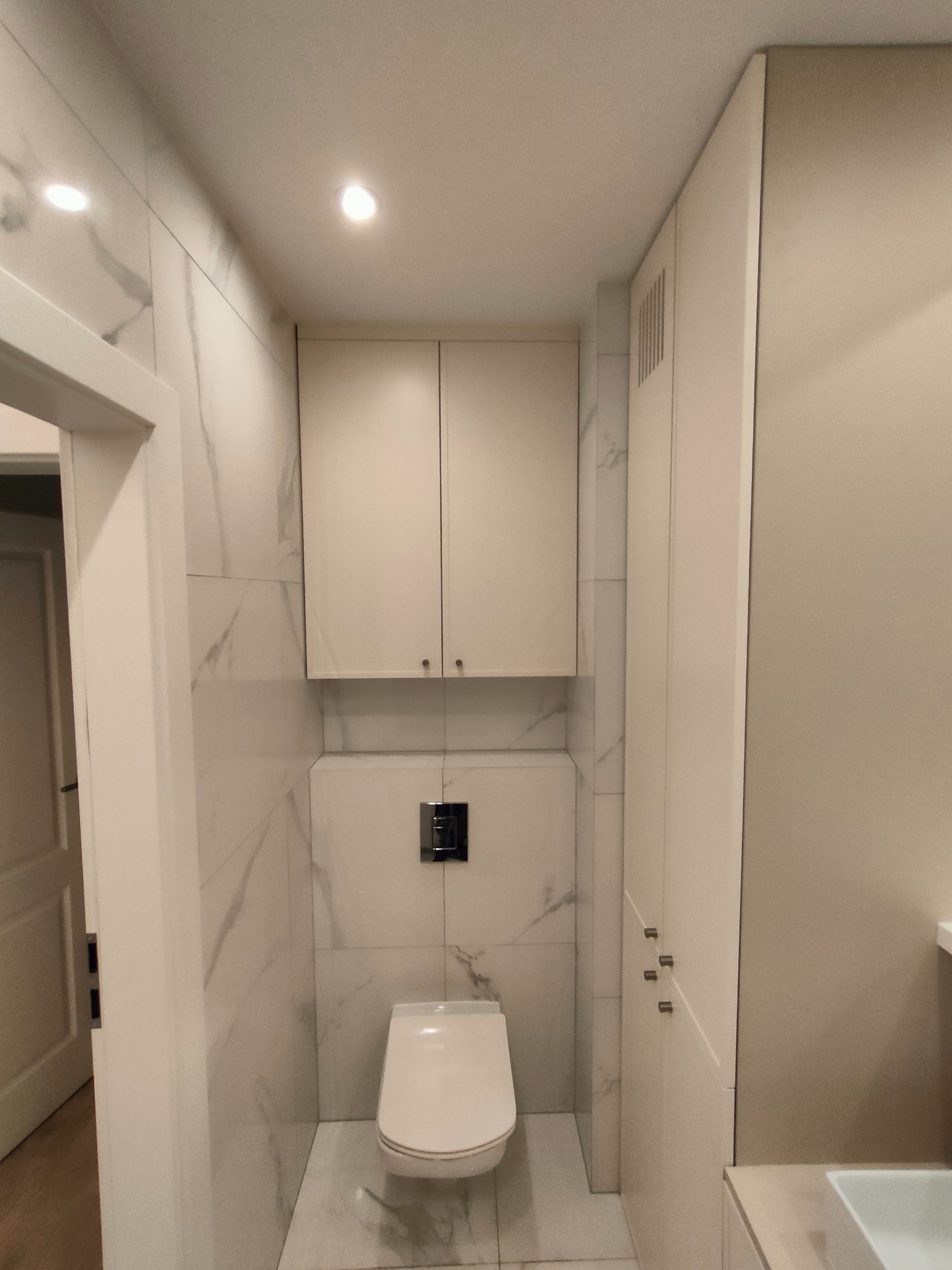 Nowoczesna toaleta z zabudową meblową w odcieniach bieli i beżu, wykończona marmurowymi płytkami. Minimalistyczny design, oświetlenie punktowe.