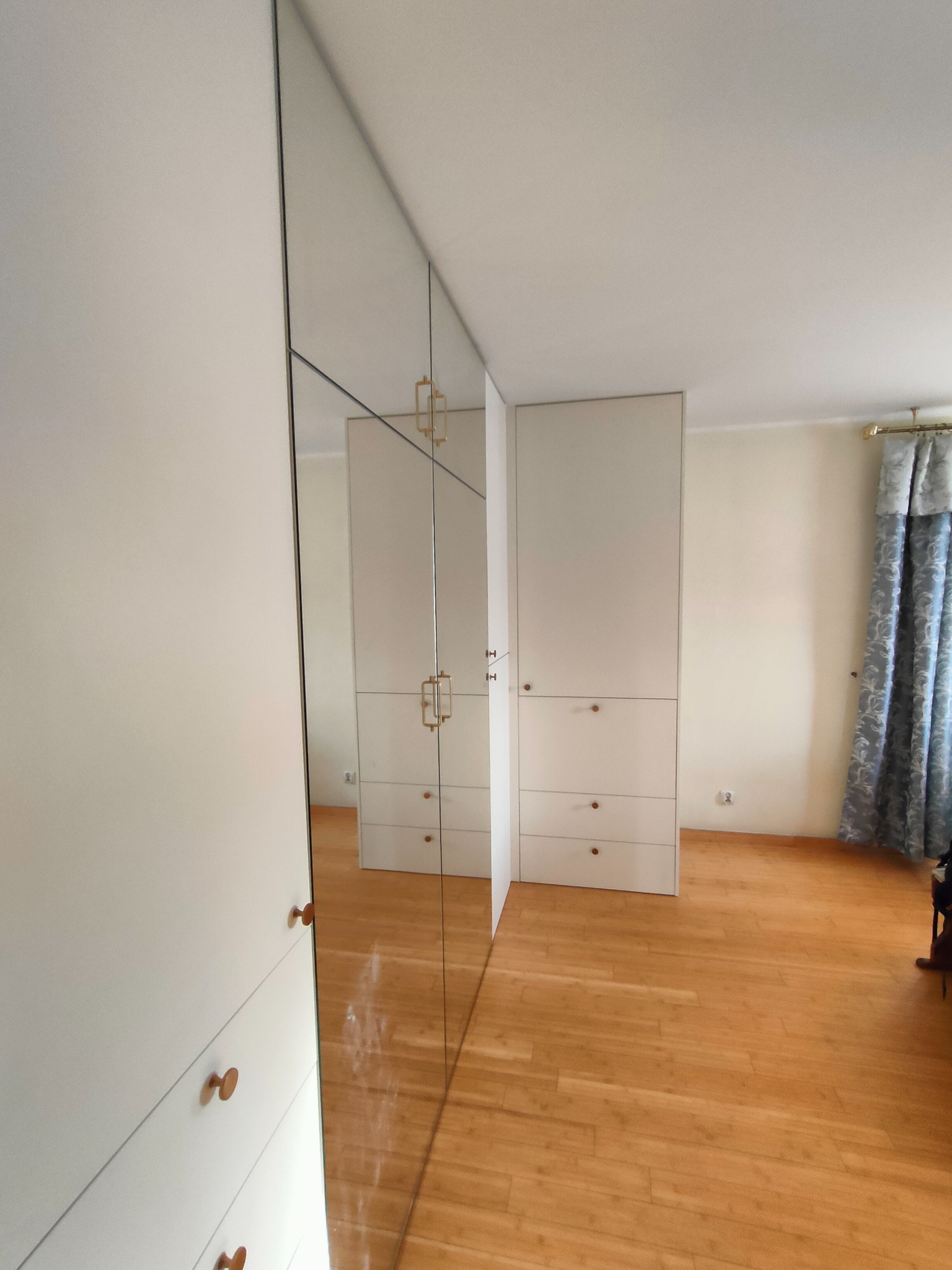 Biała szafa wnękowa z lustrzanymi drzwiami, odbijająca wnętrze pokoju z parkietem i zasłonami. Minimalistyczny design z okrągłymi uchwytami.