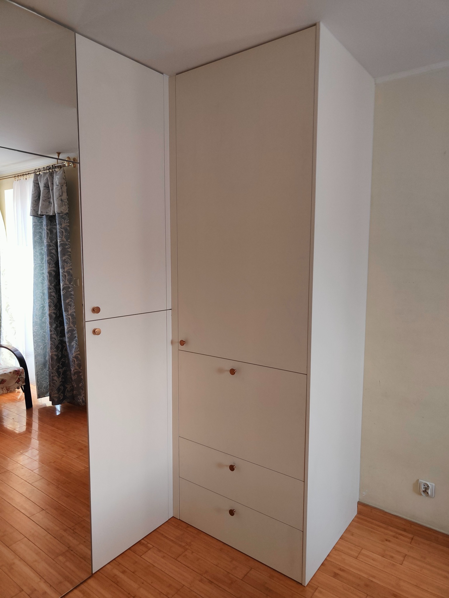 Biała szafa wnękowa z lustrzanymi drzwiami i drewnianymi uchwytami, widoczna w rogu pokoju. Minimalistyczny design, jasne wnętrze odbijające się w lustrze.