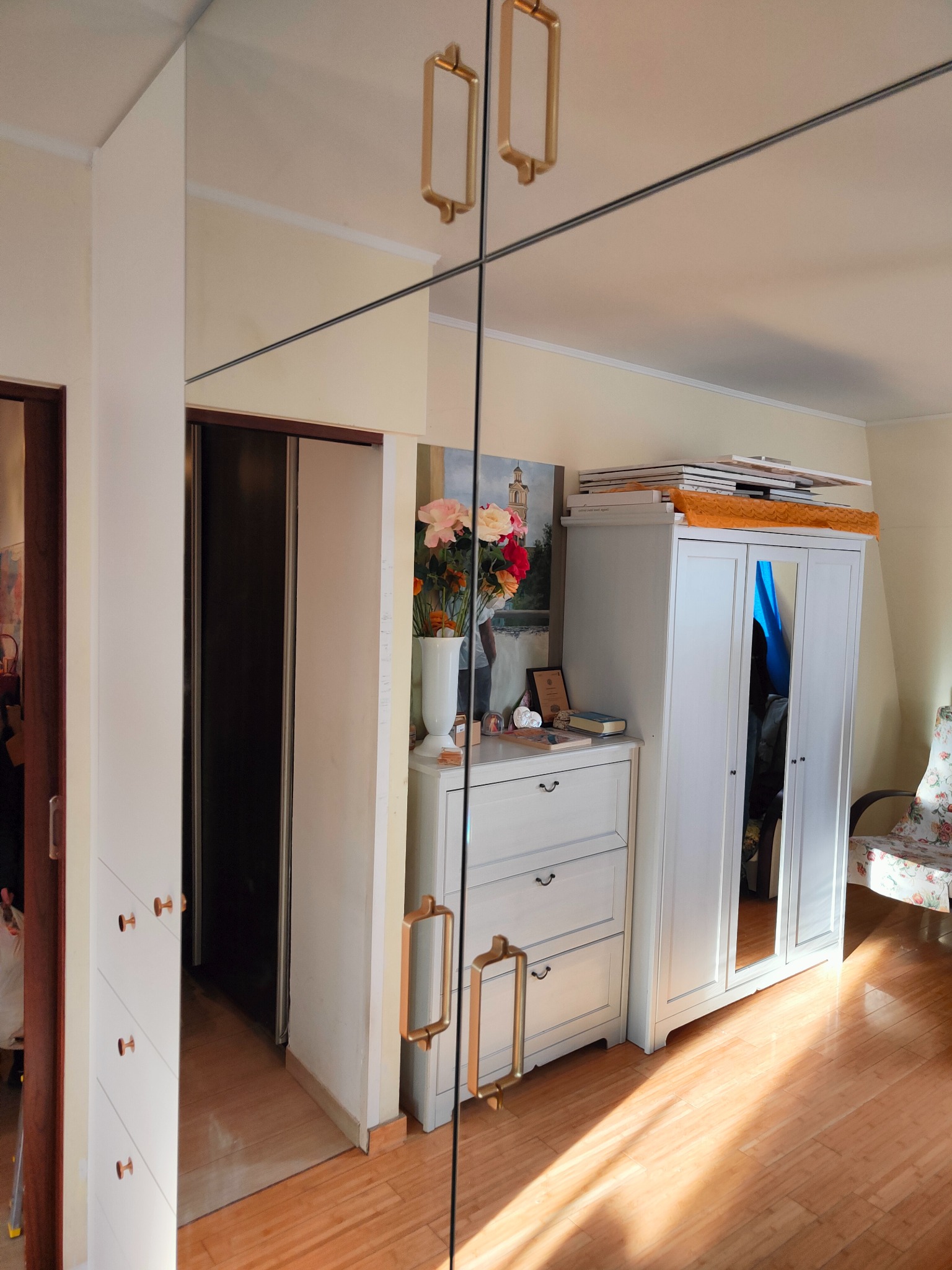 Biała szafa wnękowa z lustrzanymi drzwiami i złotymi uchwytami, odbijająca wnętrze pokoju z komodą i szafą. Widoczny parkiet i dekoracje.