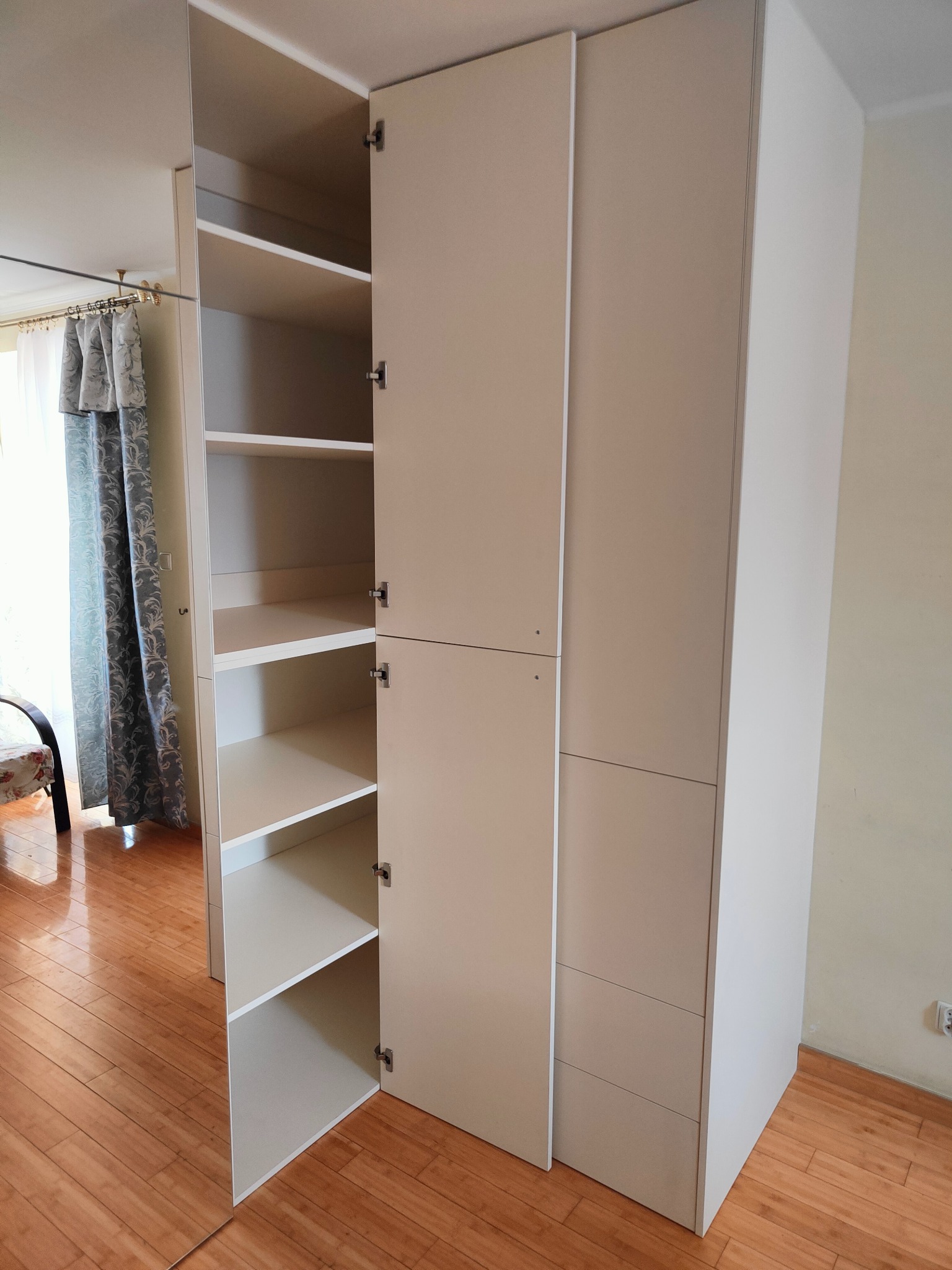 Jasna szafa wnękowa z otwartymi drzwiami, ukazująca półki. Widoczne detale wykończenia i dopasowanie do wnętrza. Na podłodze parkiet.