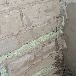 Ryszard Biłyk - Ściana z cegły wypełniona pianką montażową w szczelinach, przygotowana do dalszych prac wykończeniowych w rogu pomieszczenia. Widoczne ślady zaprawy i nierówności.