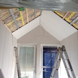 Ryszard Biłyk - Montaż płyt karton-gips na poddaszu ze skosami, widoczne okna i drabina. Izolacja folią paroizolacyjną. Przewody elektryczne zwisają z sufitu.