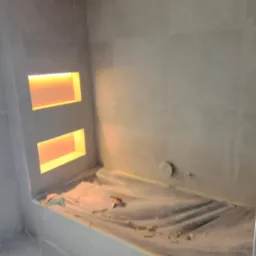Wykończona wnęka z oświetleniem w szarej, kafelkowanej łazience, zabezpieczona folią malarską. Widoczne narzędzia glazurnicze.