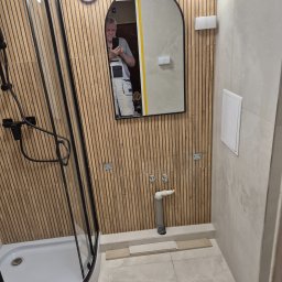 Ryszard Biłyk - Nowoczesna łazienka w trakcie wykańczania: kabina prysznicowa, lustro z odbiciem osoby, drewniane panele ścienne i jasne płytki podłogowe. Widoczne elementy instalacji.