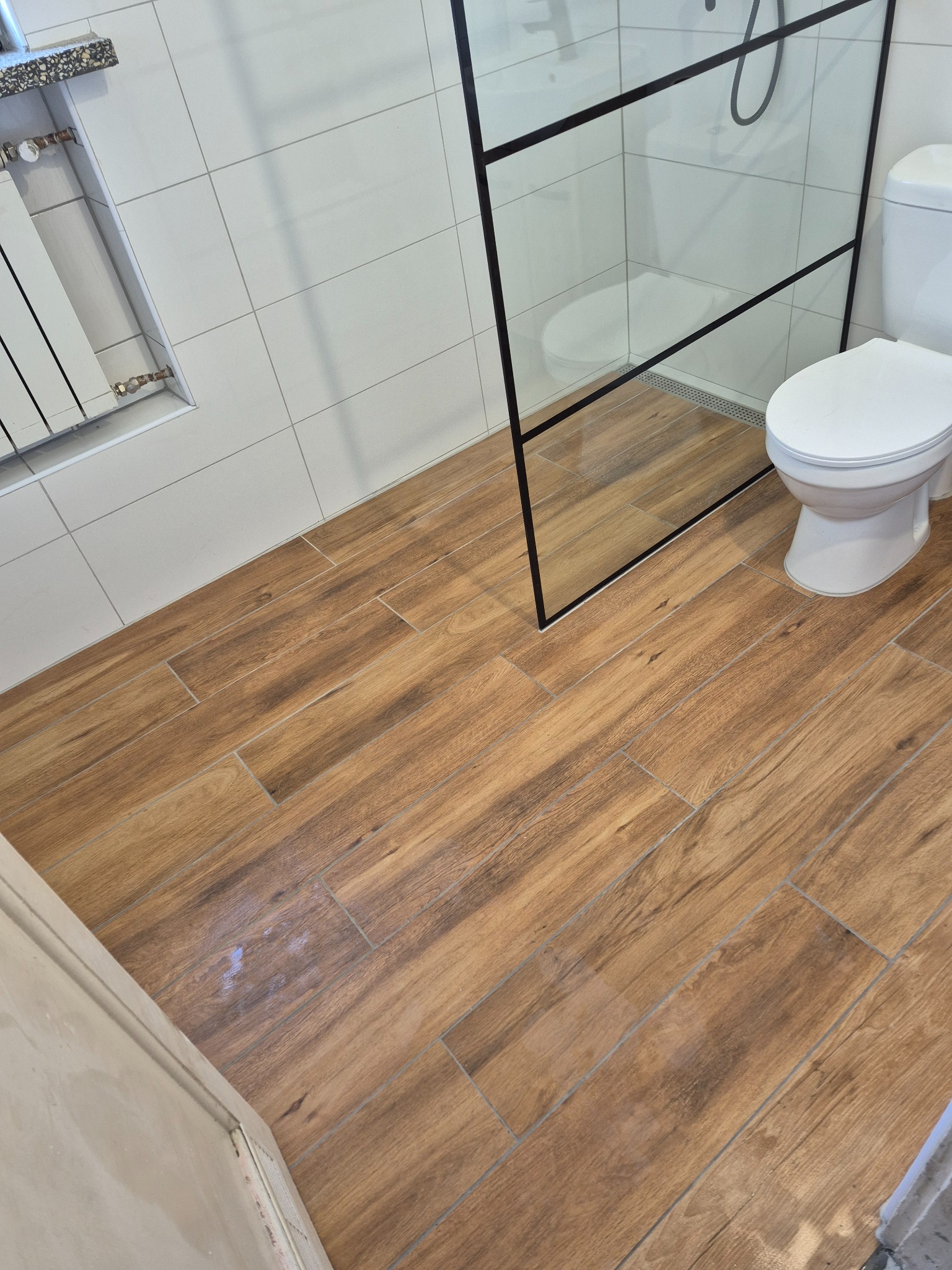 Wykończona łazienka z drewnopodobną podłogą, kabiną prysznicową typu walk-in z czarnymi ramami i białą ceramiką. Grzejnik drabinkowy na ścianie z białych płytek.