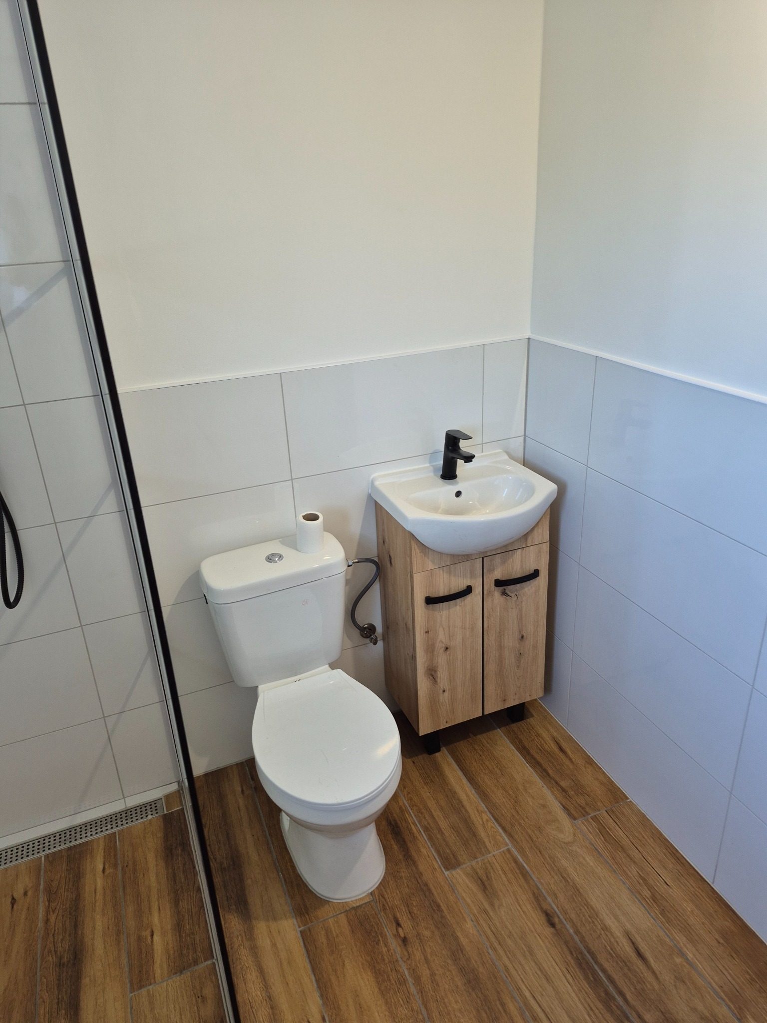 Nowoczesna łazienka po remoncie: białe płytki, drewnopodobna podłoga, podwieszana szafka z umywalką i czarną armaturą, kompakt WC z podajnikiem na papier.