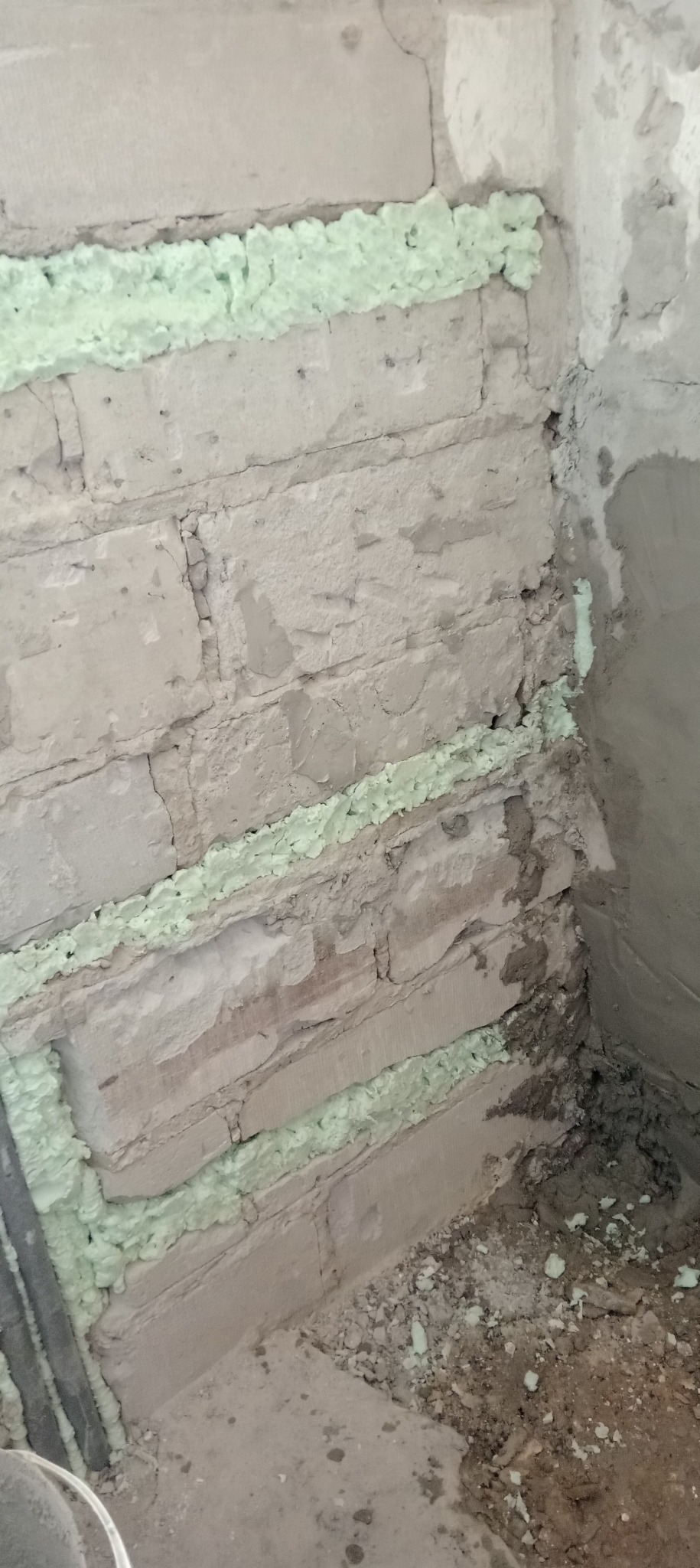 Ściana z cegły wypełniona pianką montażową w szczelinach, przygotowana do dalszych prac wykończeniowych w rogu pomieszczenia. Widoczne ślady zaprawy i nierówności.
