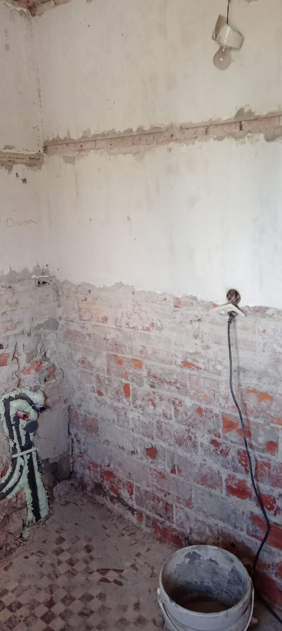 Ceglana ściana w trakcie prac remontowych, częściowo otynkowana, z widoczną instalacją i wiszącą żarówką. Na podłodze resztki gruzu i wiadro.