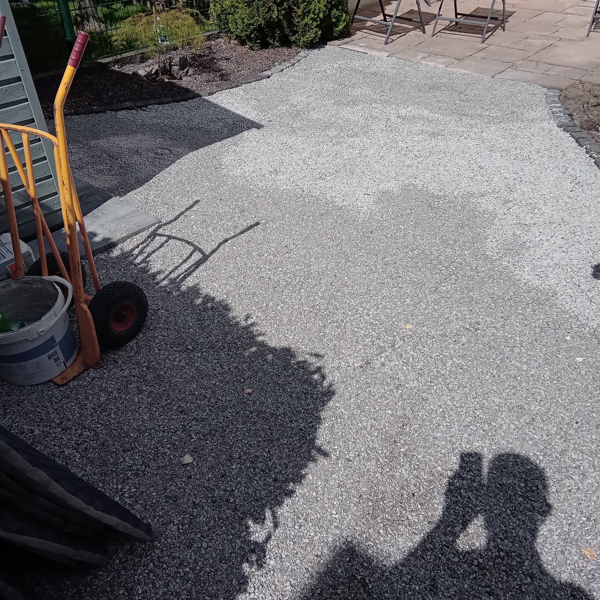 Świeżo wysypany żwir na podwórku, widoczny wózek z narzędziami i cień fotografa. Granica z kostki brukowej oddziela żwir od trawnika i płyt chodnikowych.