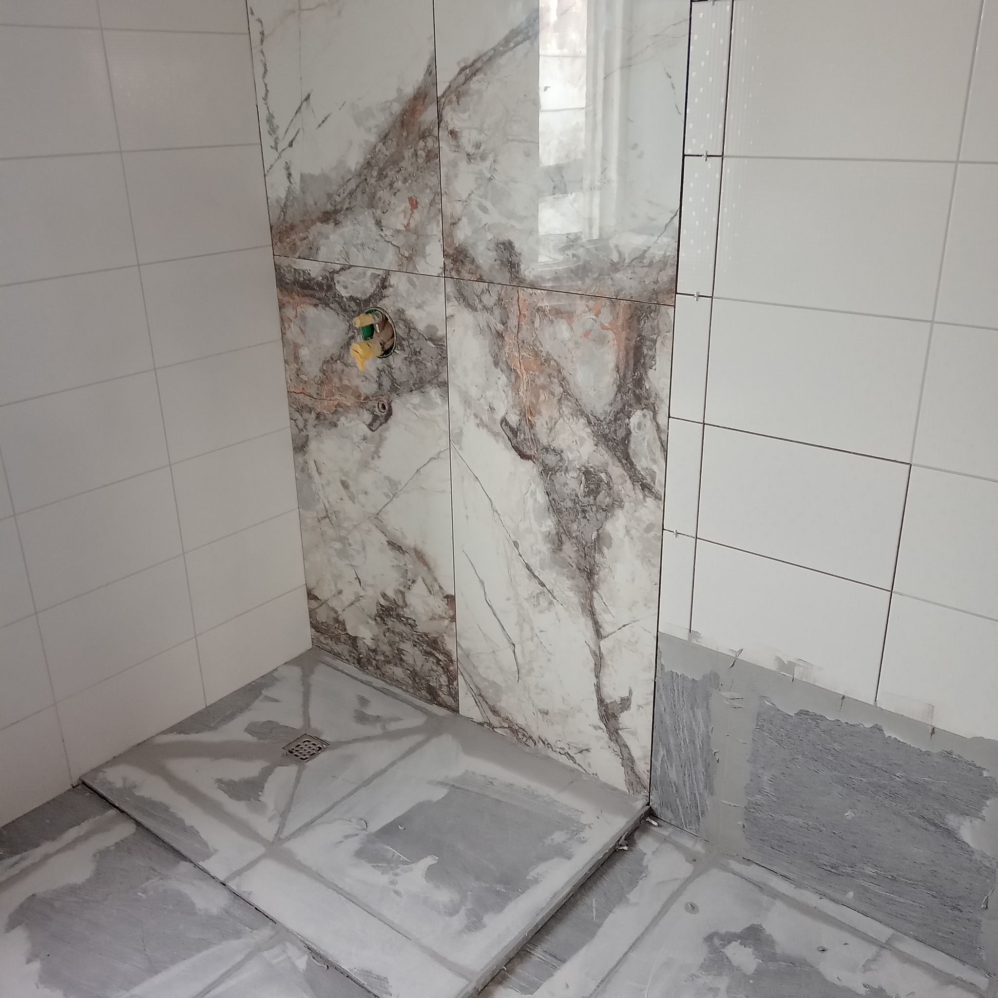 Fragment łazienki w trakcie prac. Ułożone białe i marmurowe płytki na ścianach. Uformowany brodzik z odpływem. Widoczne ślady zaprawy.