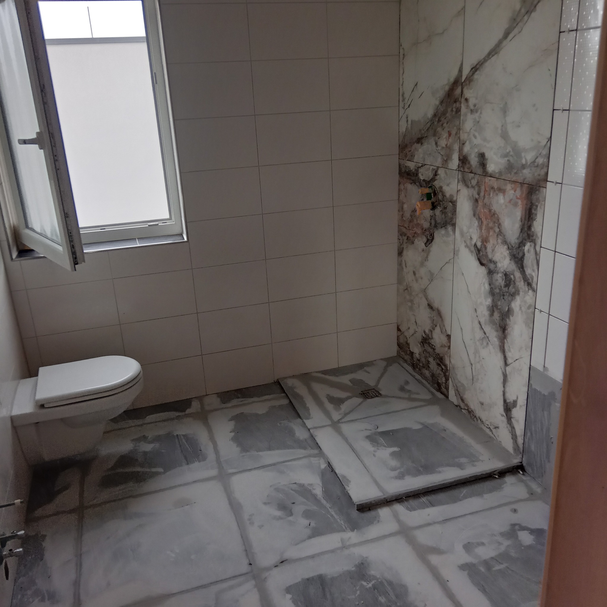 Łazienka w trakcie remontu: białe płytki na ścianach, marmurowe akcenty w strefie prysznica, częściowo ułożona podłoga, toaleta pod oknem. Widoczny etap prac glazurniczych.