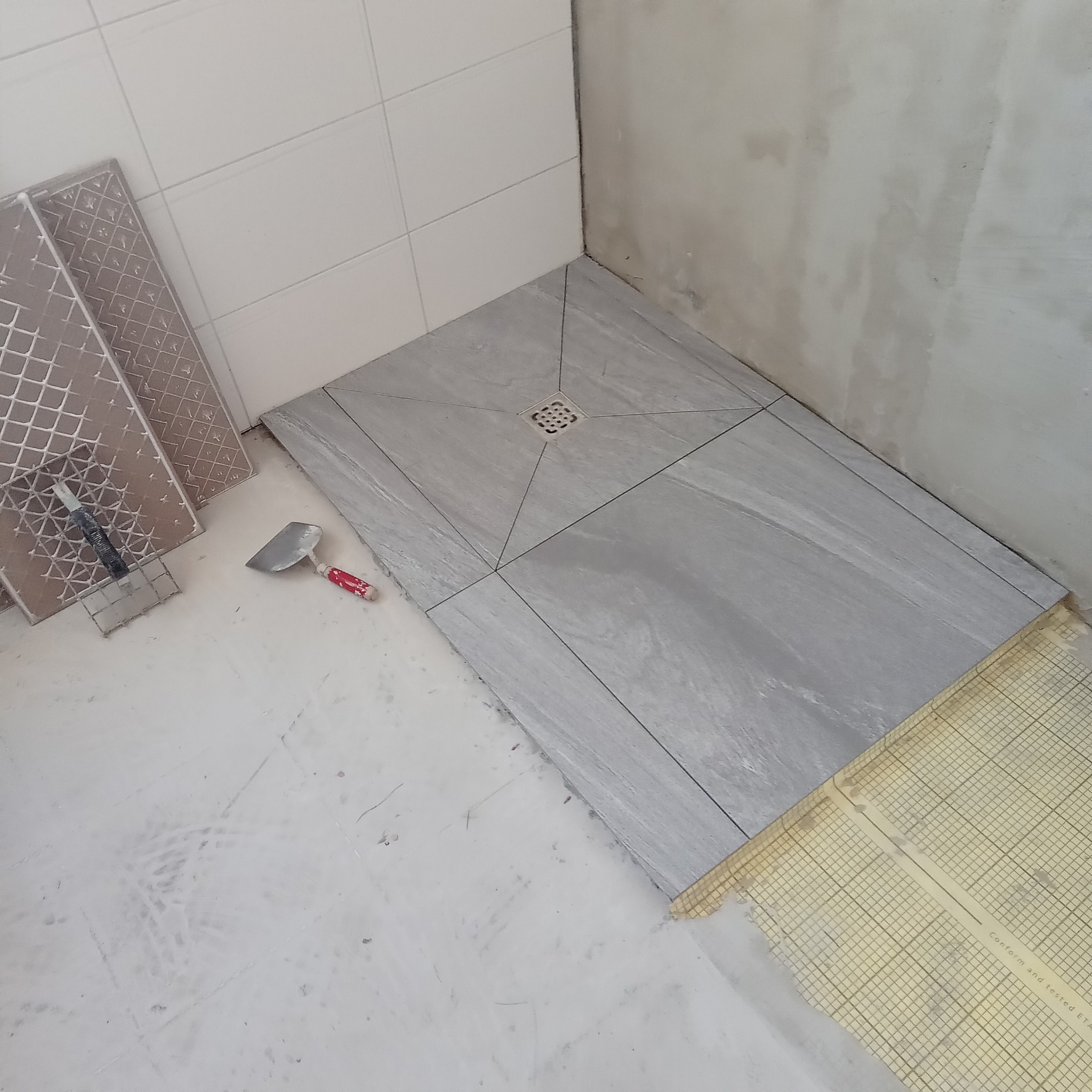 Układanie szarych płytek podłogowych w łazience, widoczny odpływ liniowy i narzędzia glazurnicze. Na ścianach białe płytki i surowy beton.