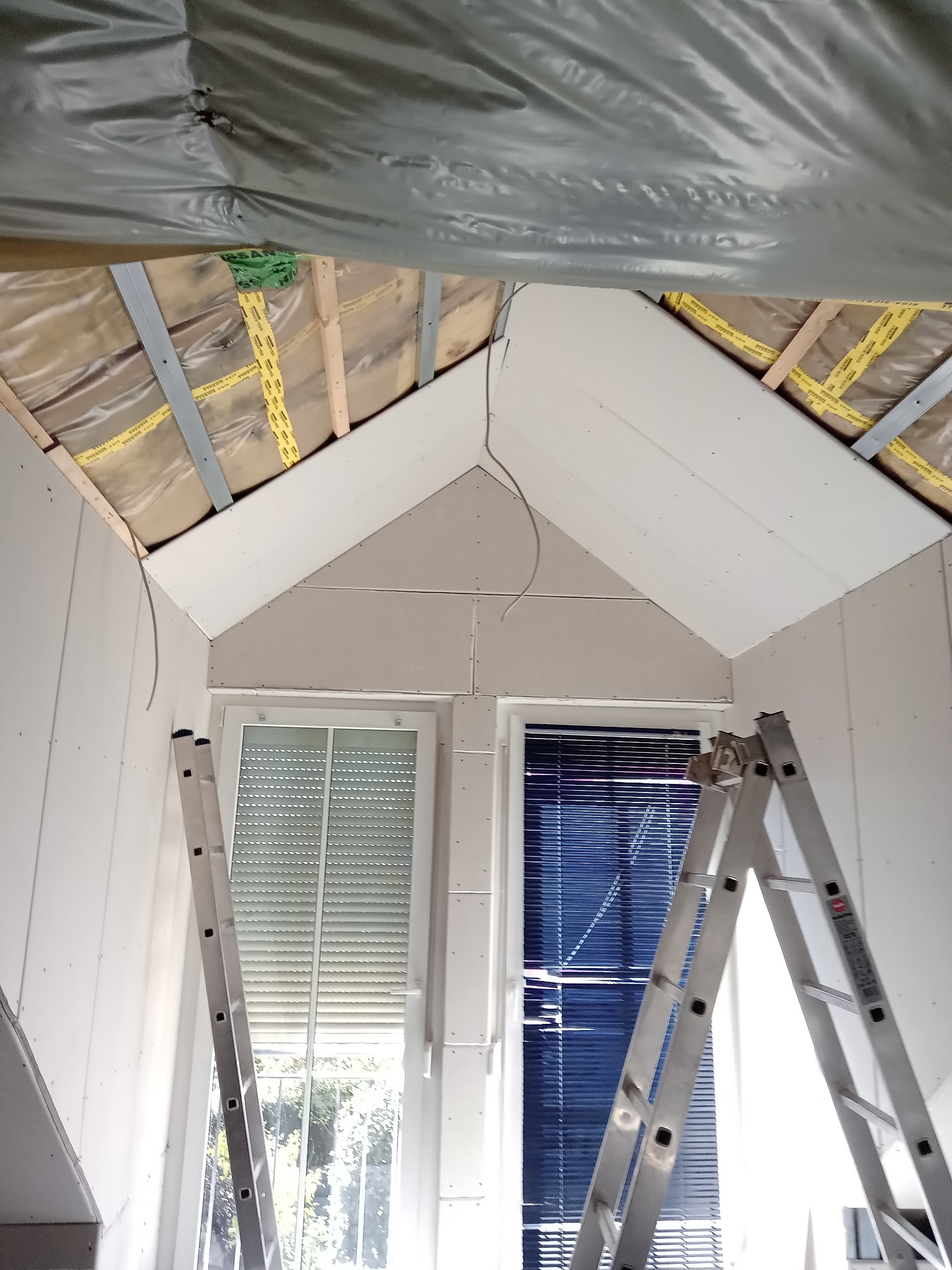 Montaż płyt karton-gips na poddaszu ze skosami, widoczne okna i drabina. Izolacja folią paroizolacyjną. Przewody elektryczne zwisają z sufitu.