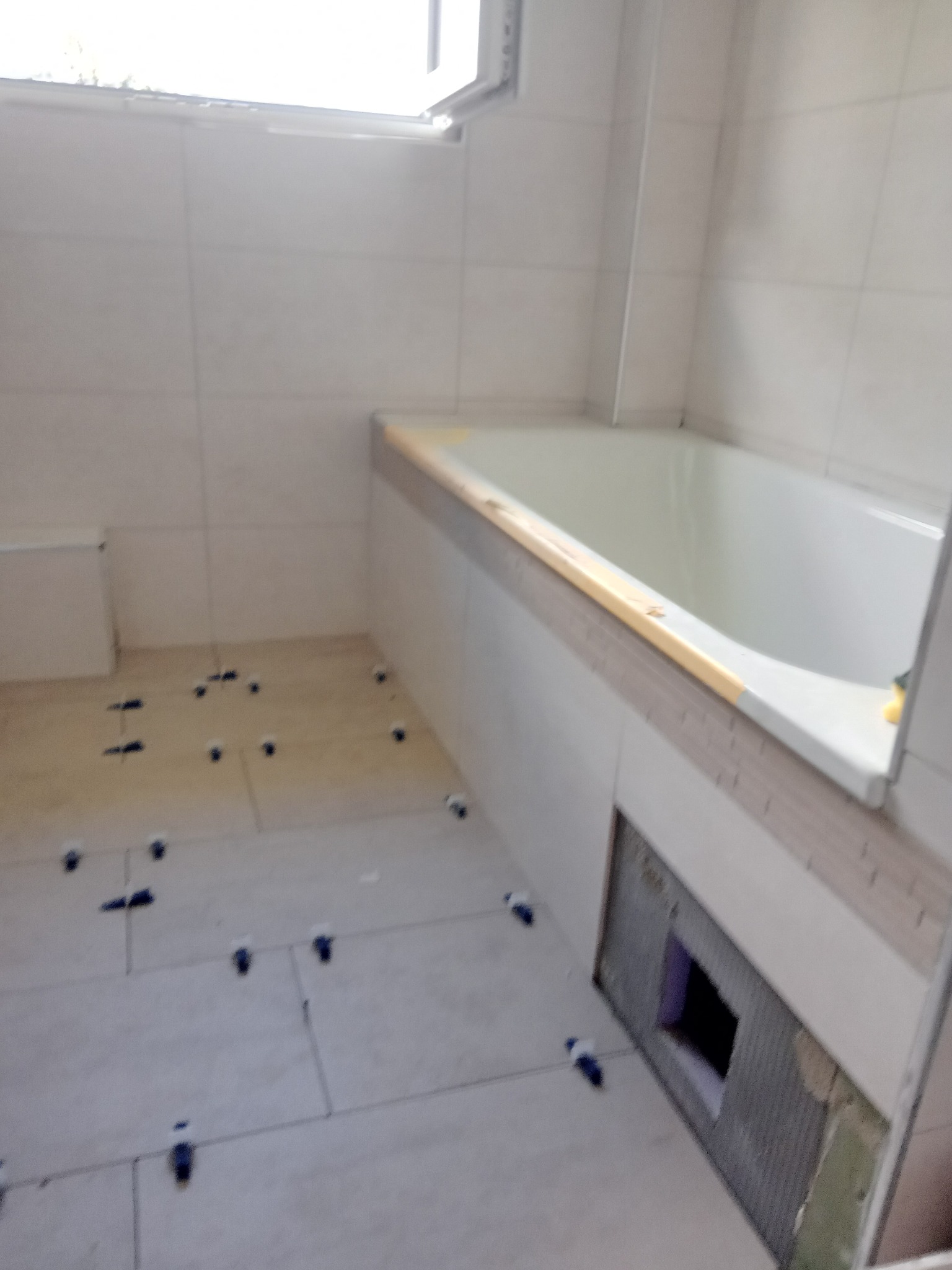 Łazienka w trakcie układania płytek podłogowych, widoczna wanna i okno. Użyto klipsów poziomujących. Otwór rewizyjny w zabudowie wanny.