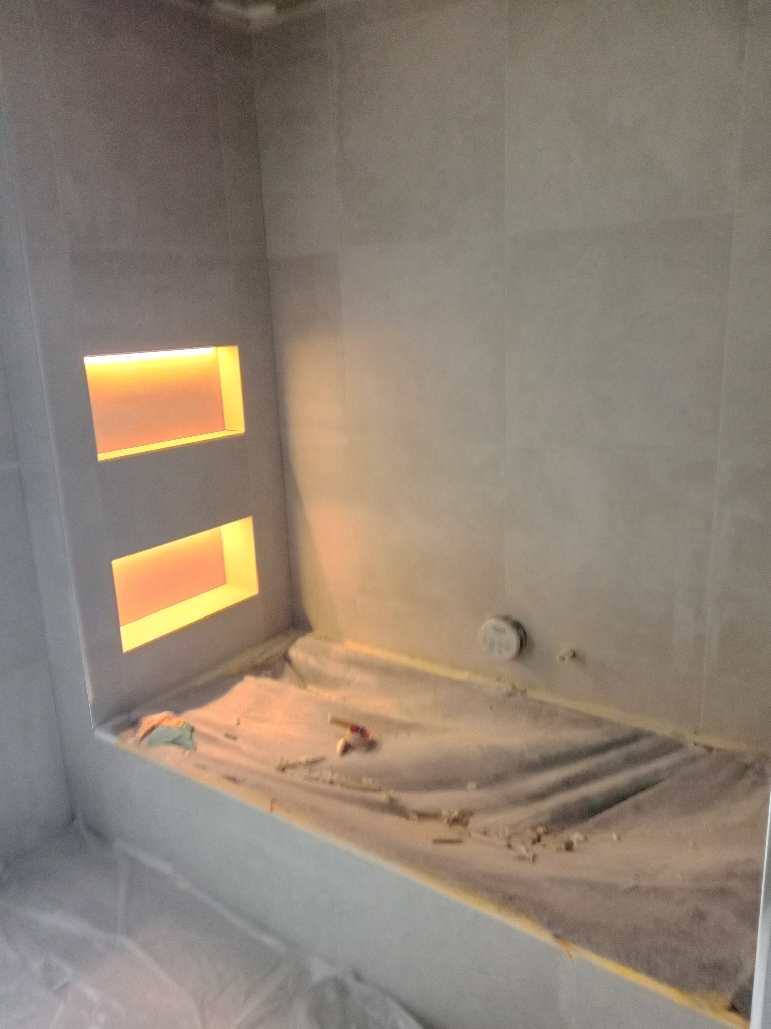 Wykończona wnęka z oświetleniem w szarej, kafelkowanej łazience, zabezpieczona folią malarską. Widoczne narzędzia glazurnicze.