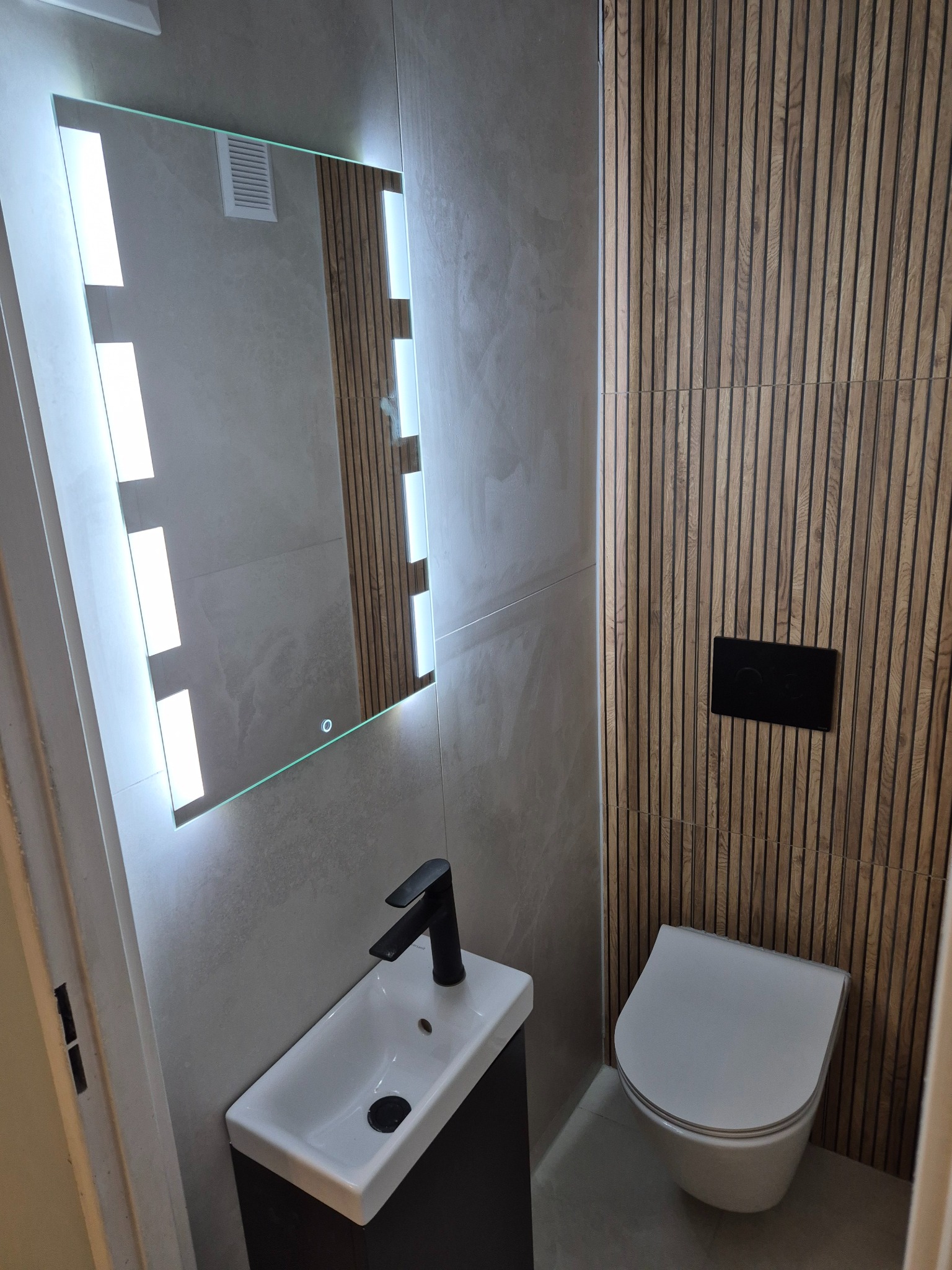 Nowoczesna łazienka z podświetlanym lustrem, drewnianą ścianą z lameli i minimalistyczną armaturą. Umywalka z czarną baterią, wisząca toaleta. Stonowana kolorystyka.