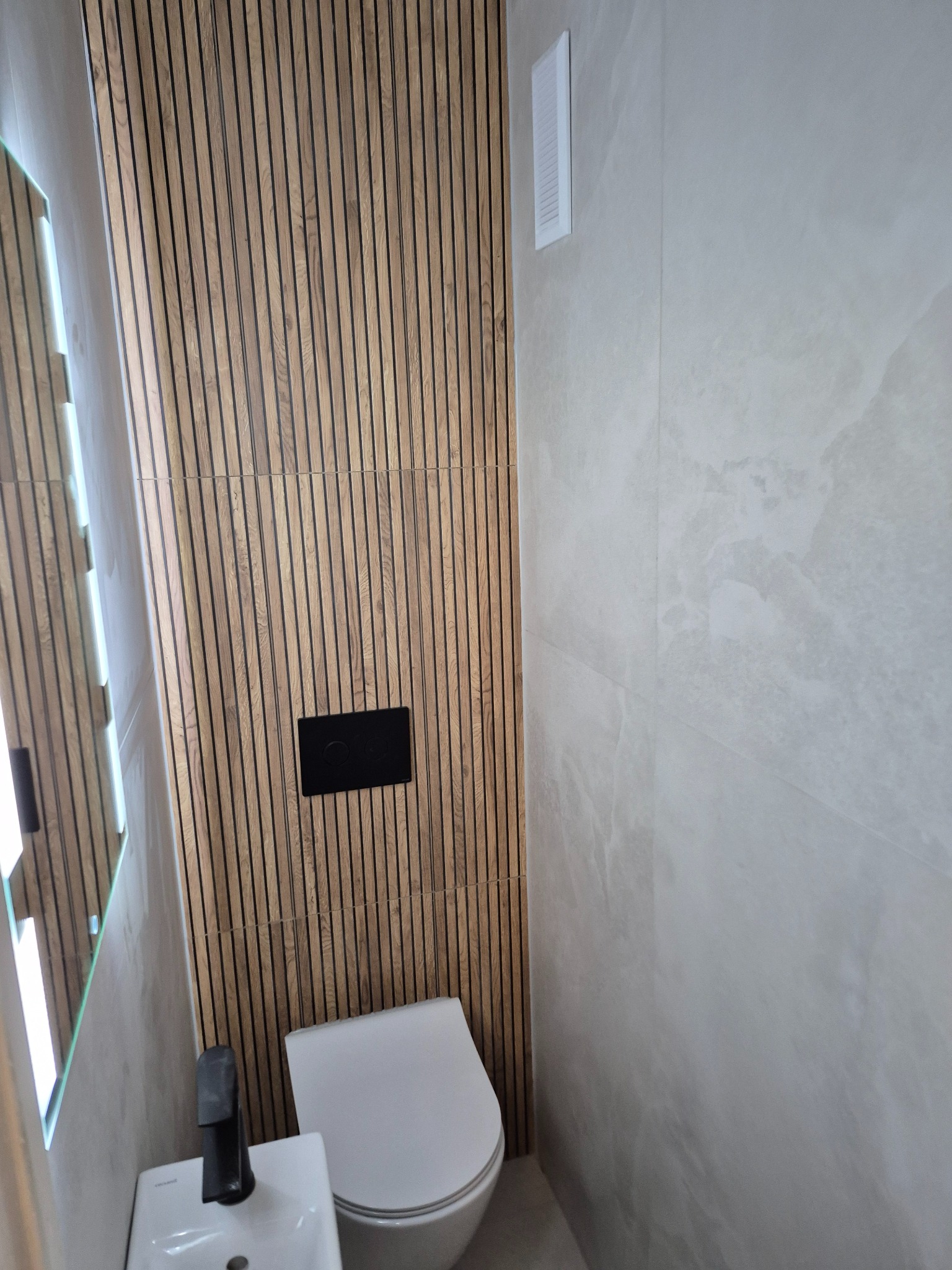 Nowoczesna łazienka z drewnianą ścianą z lameli, umywalką z czarną armaturą i podwieszaną toaletą. Minimalistyczny design, jasne płytki imitujące marmur.