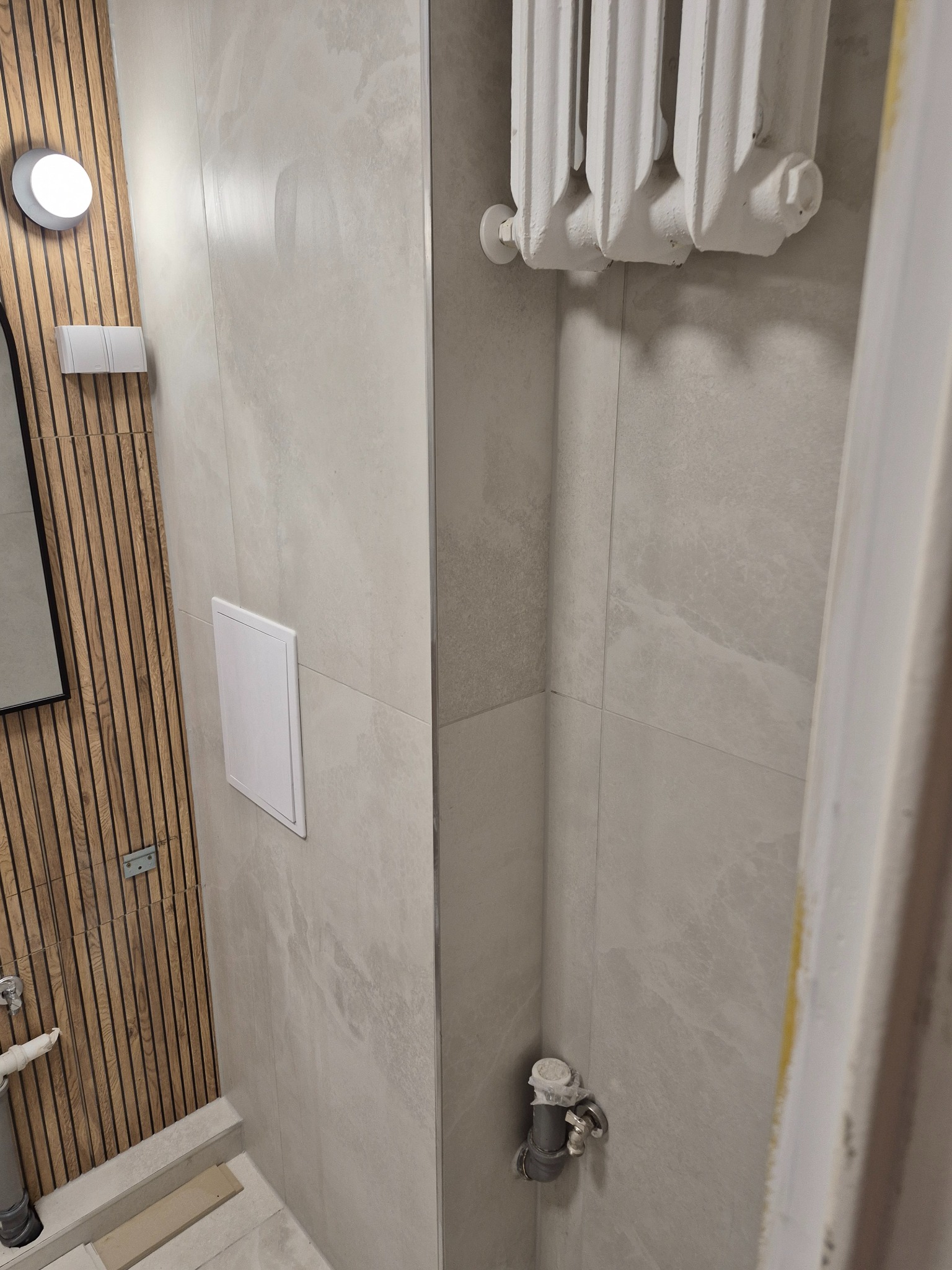 Wykończona łazienka z drewnianą ścianą, szarą płytką imitującą kamień, białym grzejnikiem i ukrytym zaworem rewizyjnym. Minimalistyczny design, nowoczesne wykończenie.