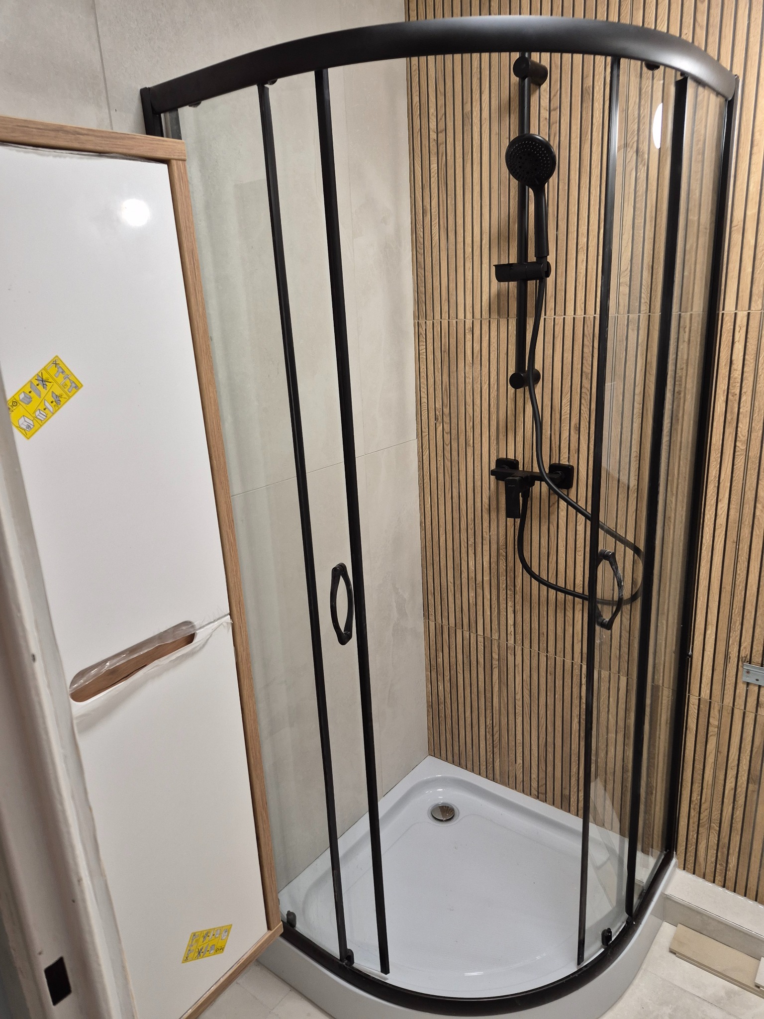 Nowoczesna kabina prysznicowa z czarnymi profilami i deszczownicą na tle drewnopodobnych paneli ściennych. Częściowo widoczna biała szafka z instrukcją montażu.