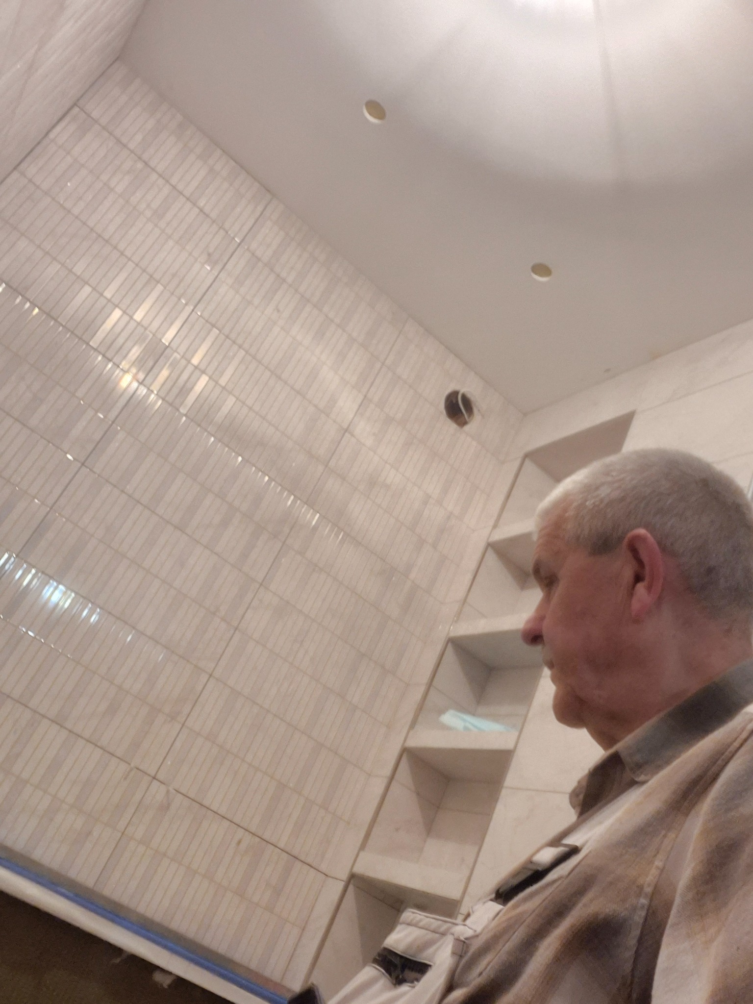 Wykończona łazienka z beżowymi, błyszczącymi płytkami ułożonymi pionowo. Widoczny fragment sufitu z oświetleniem i osoba w prawym dolnym rogu kadru.