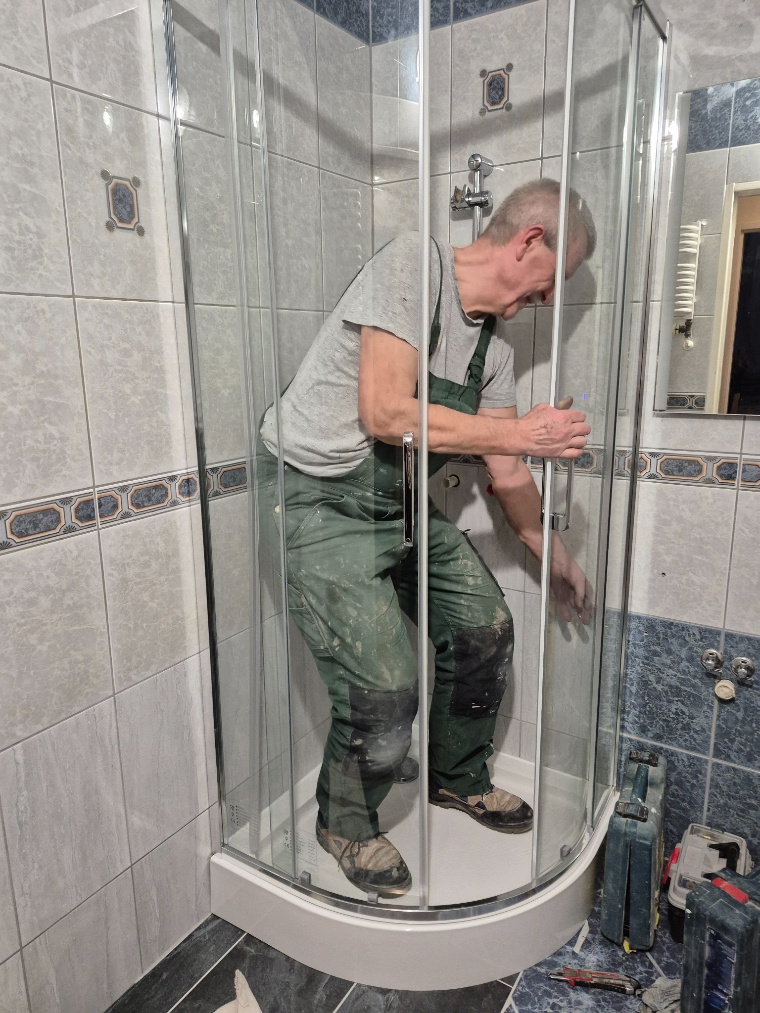 Instalacja kabiny prysznicowej w łazience z szarymi płytkami. Fachowiec w zielonym kombinezonie montuje drzwi kabiny. Widoczne narzędzia i skrzynki narzędziowe.
