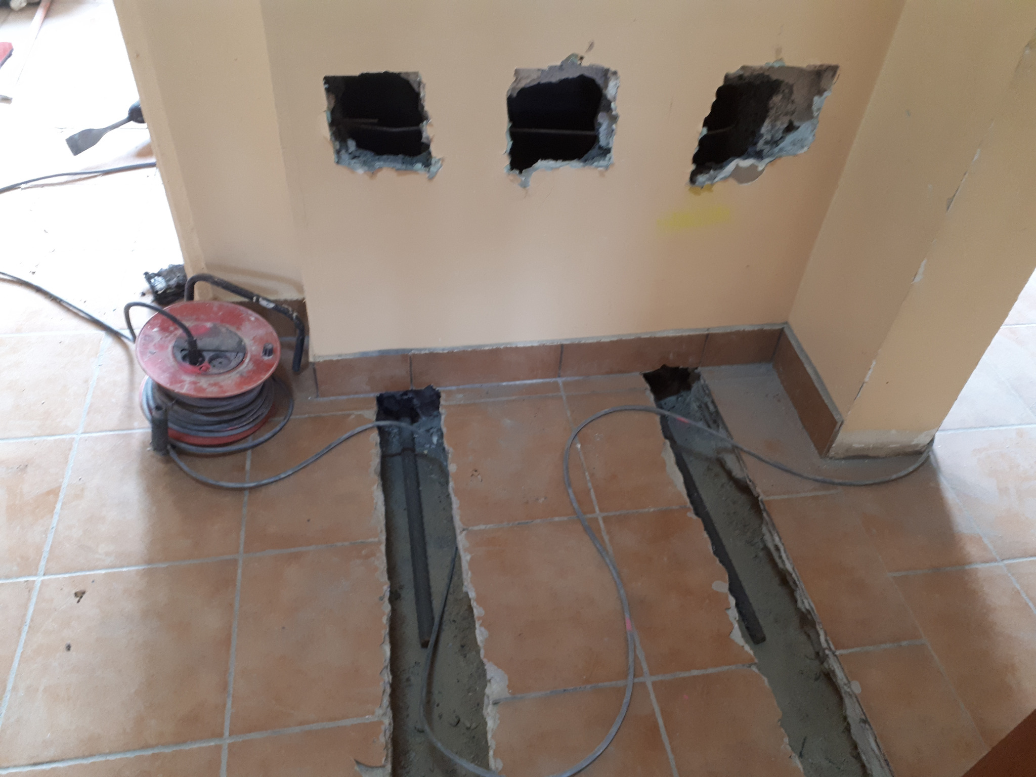 Przygotowanie do układania płytek: Wykute bruzdy w podłodze i otwory w ścianie, widoczna zwinięta przedłużka elektryczna i fragment ściany z usuniętymi płytkami.