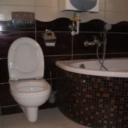 Nowoczesna łazienka z toaletą z otwartą klapą, narożną wanną obłożoną mozaiką, brązowymi płytkami na ścianie z dekoracyjnym wzorem oraz podgrzewaczem wody nad wanną.
