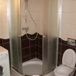 Narożna kabina prysznicowa z matowymi szybami w łazience z brązowo-białymi płytkami i toaletą, obok pralka.