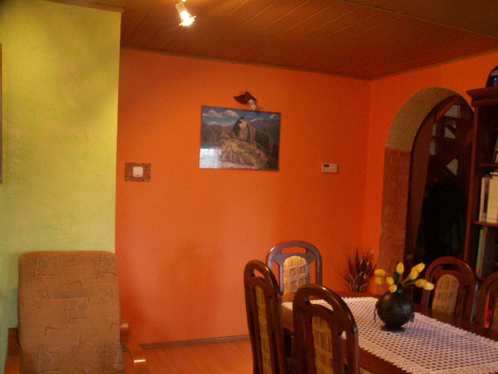 Pomarańczowa ściana w jadalni z obrazem górskim, zielona ściana po lewej, drewniane krzesła z żółtymi akcentami i stół przykryty białym obrusem z koronką, na stole wazon z żółtymi tulipanami.