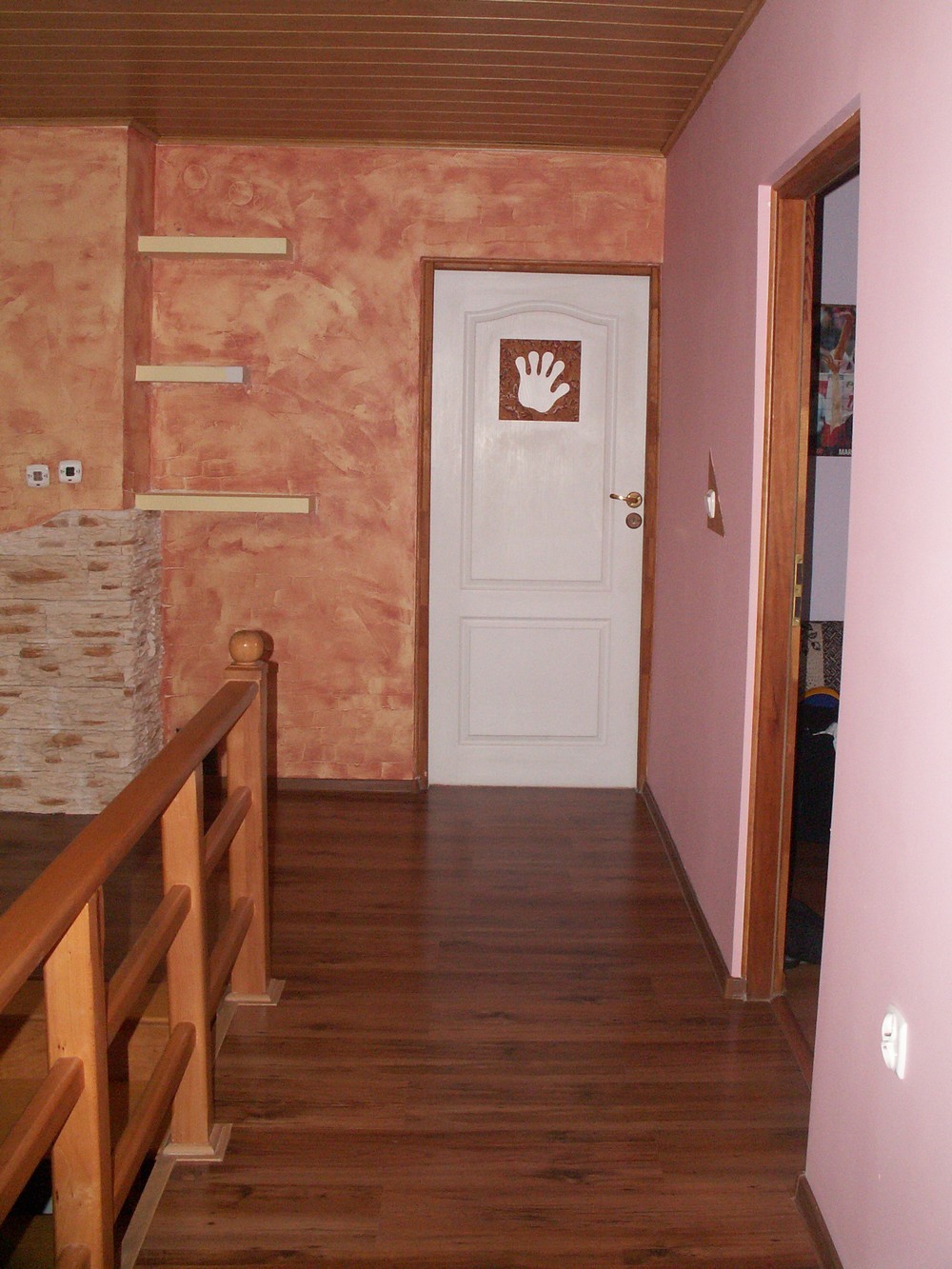 Korytarz po remoncie z drewnianą podłogą, ścianą w odcieniach pomarańczy z półkami i białą wewnętrzną stolarką drzwiową z naklejoną grafiką dłoni.