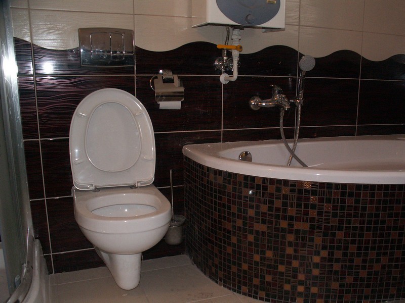Nowoczesna łazienka z toaletą z otwartą klapą, narożną wanną obłożoną mozaiką, brązowymi płytkami na ścianie z dekoracyjnym wzorem oraz podgrzewaczem wody nad wanną.