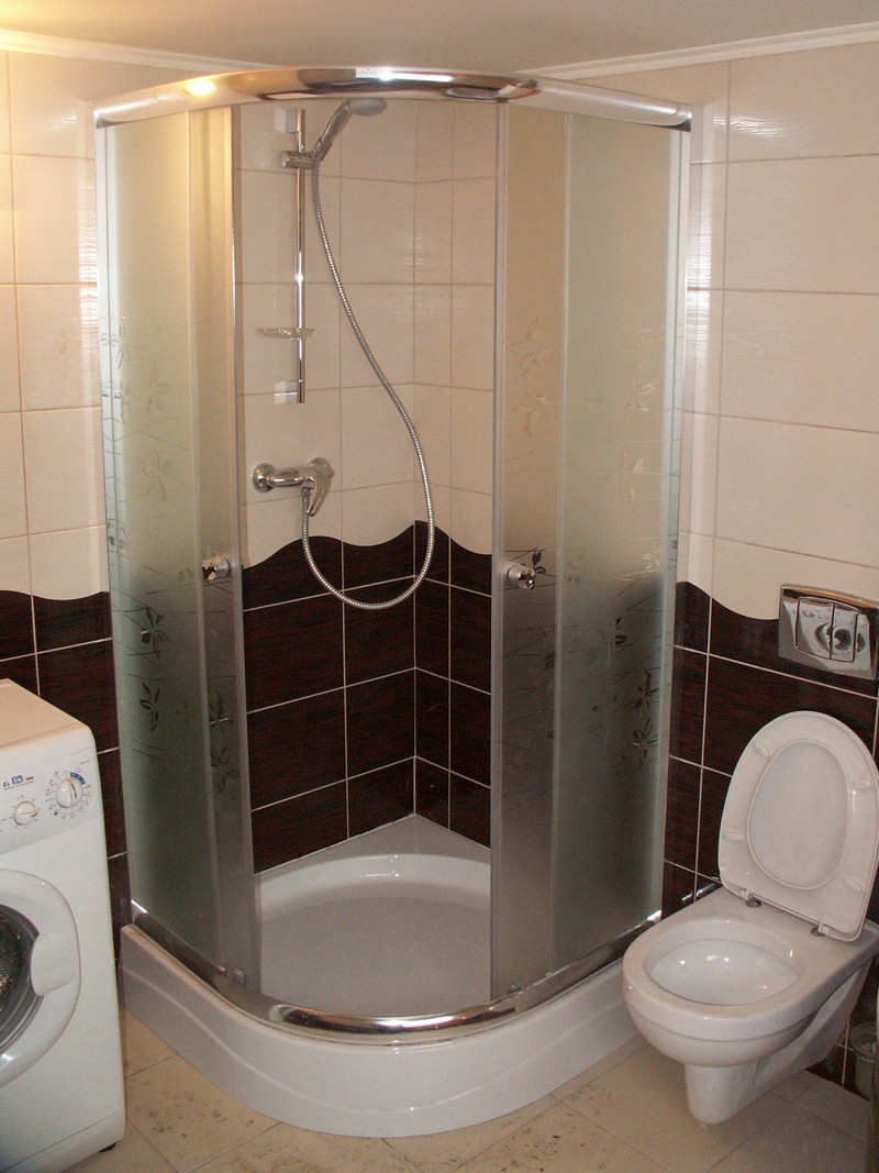 Narożna kabina prysznicowa z matowymi szybami w łazience z brązowo-białymi płytkami i toaletą, obok pralka.