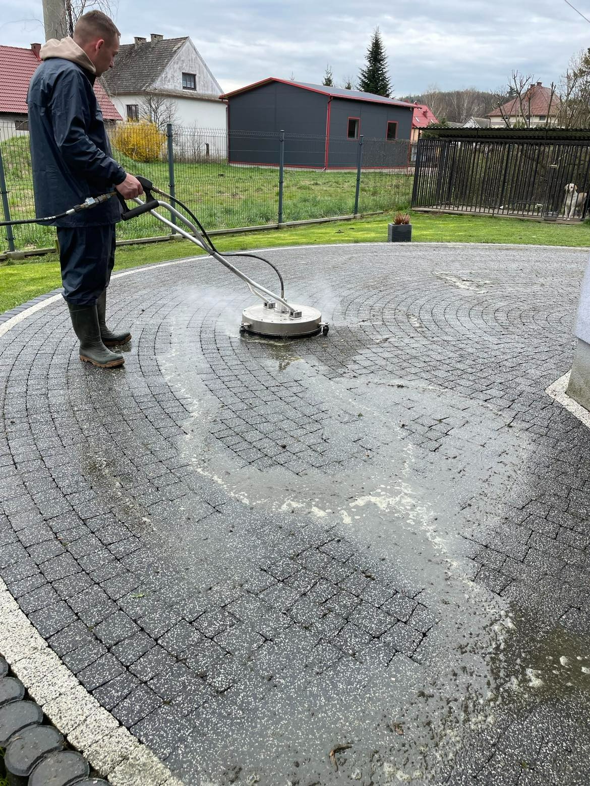 Mężczyzna w kaloszach czyści okrągły podjazd z kostki brukowej za pomocą myjki ciśnieniowej z okrągłą dyszą; widoczne zabrudzenia i spływająca woda.