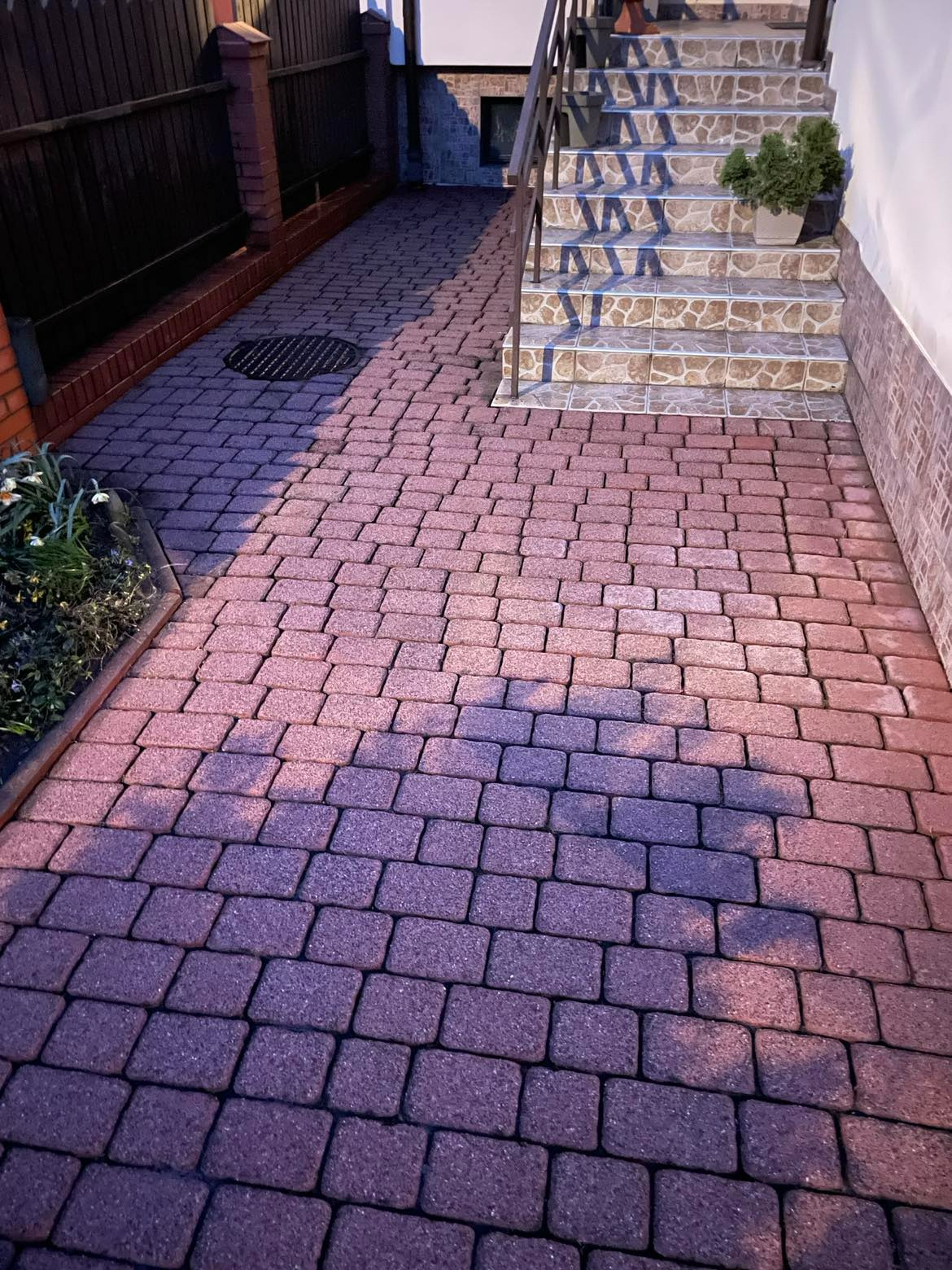 Ułożona czerwono-brązowa kostka brukowa prowadząca do schodów z rośliną w doniczce, widoczna studzienka kanalizacyjna.