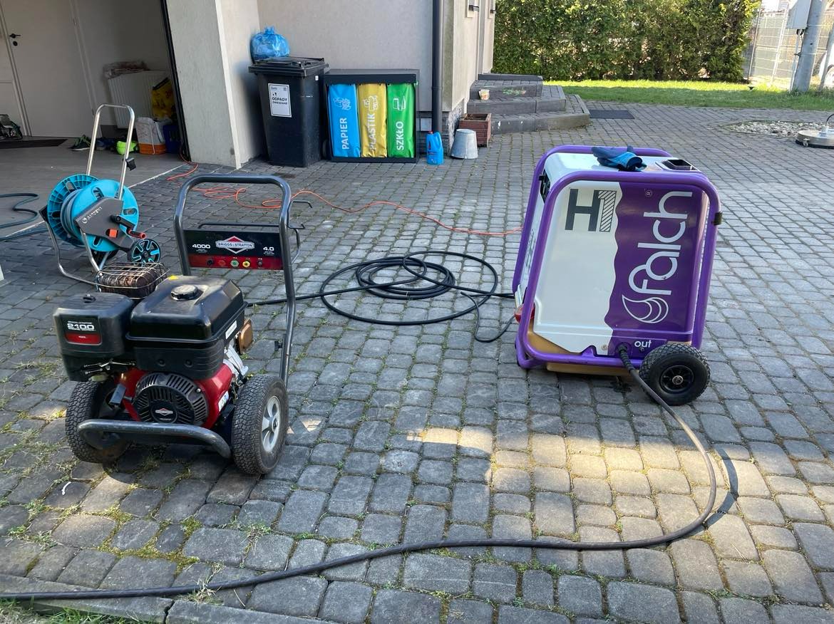 Dwa urządzenia do mycia ciśnieniowego kostki brukowej marki Falch i Briggs&Stratton, przygotowane do pracy na posesji w Krakowie, z widocznymi pojemnikami do segregacji odpadów w tle.