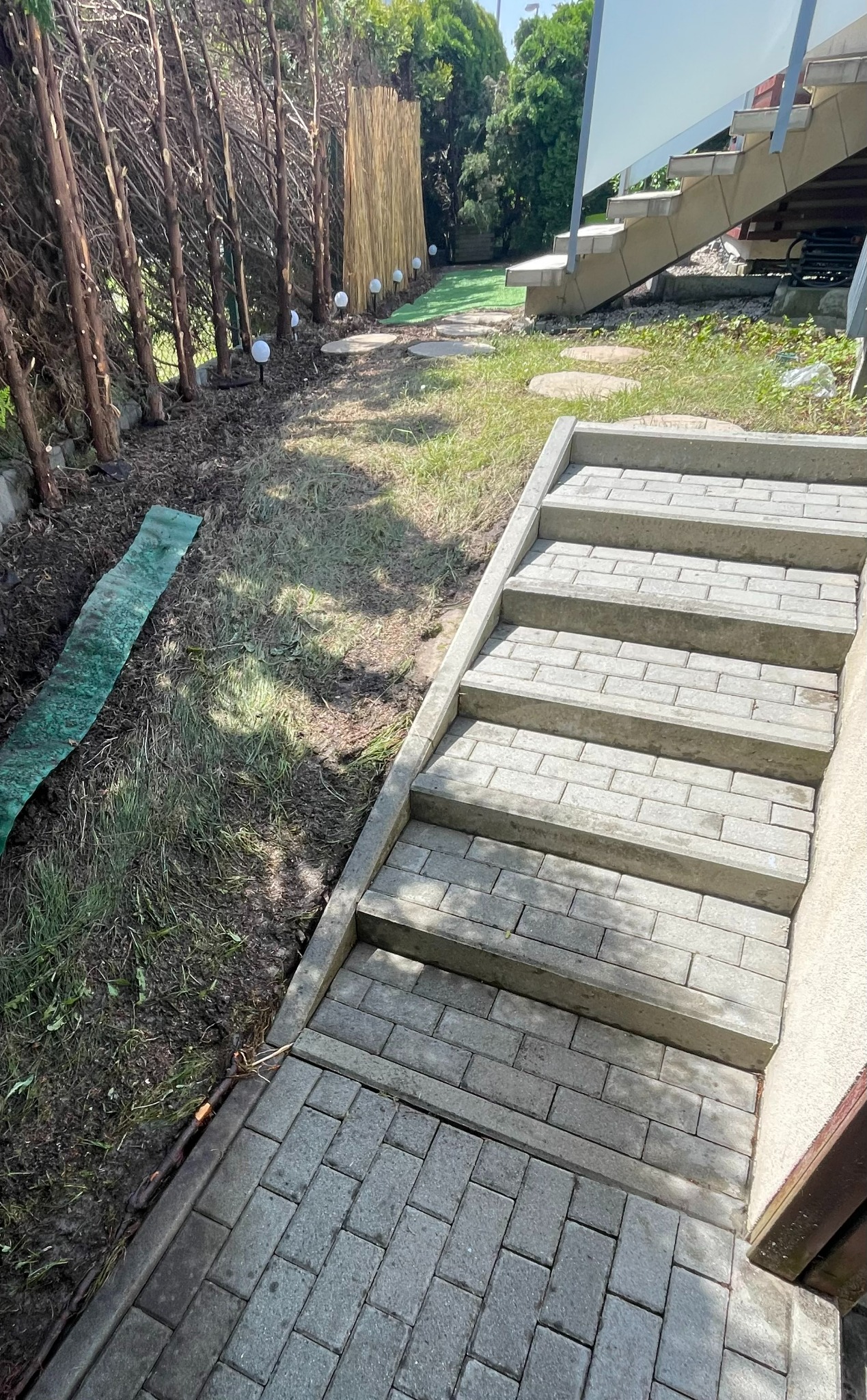 Schody z kostki brukowej prowadzące w górę, obok zaniedbany trawnik z resztkami roślinności, w tle żywopłot i elementy architektury ogrodowej.