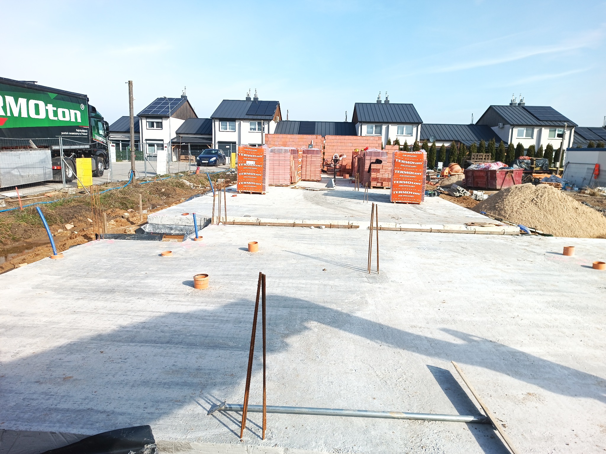 Wylany betonowy fundament pod budowę domu jednorodzinnego, widoczne wystające pręty zbrojeniowe i stosy cegieł ceramicznych Termoton w tle, na drugim planie zabudowa osiedla.