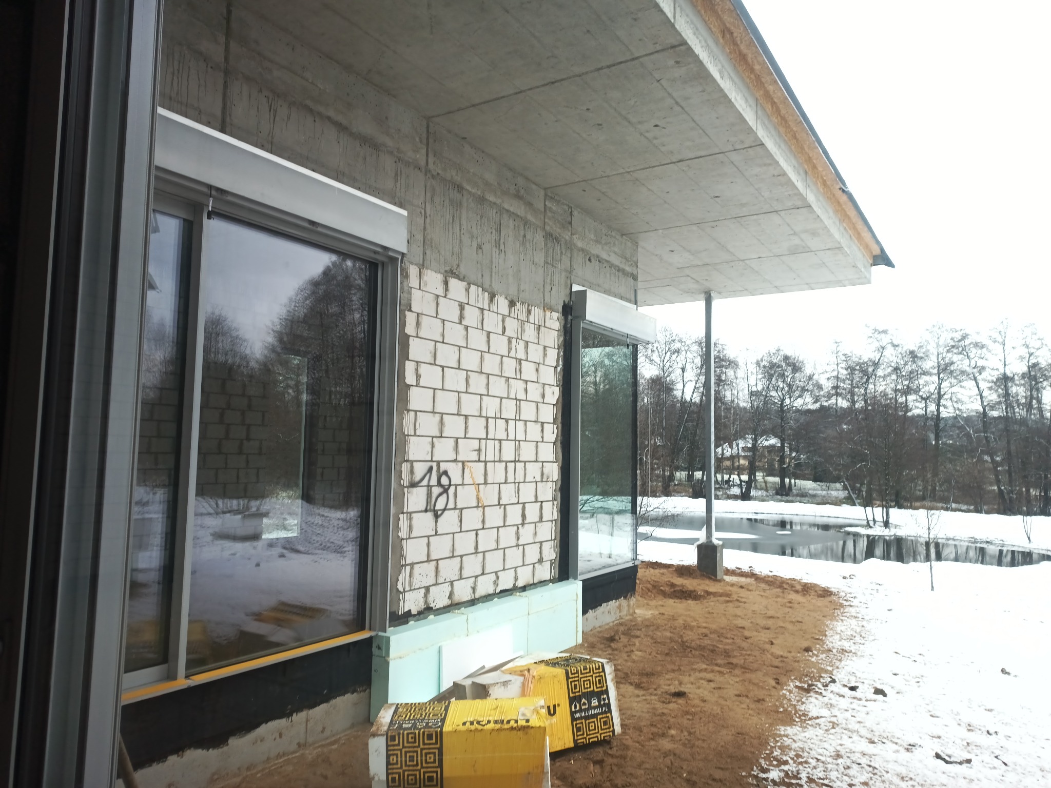 Surowy stan domu w trakcie budowy z widoczną ścianą z białej cegły, oknami z roletami zewnętrznymi, betonowym stropodachem i ociepleniem fundamentów, na tle zimowego krajobrazu z jeziorem.