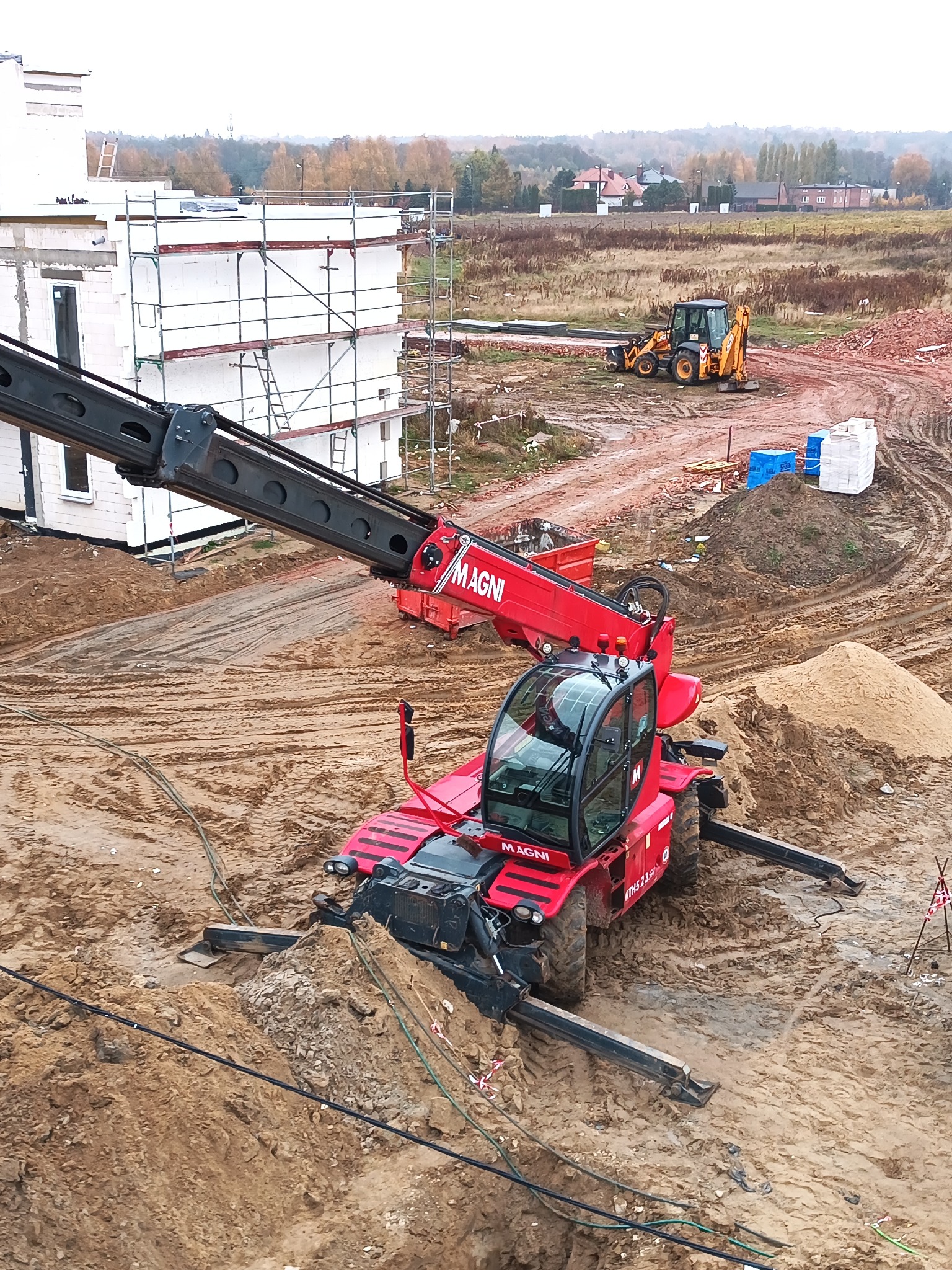 Czerwony podnośnik teleskopowy Magni na placu budowy, w tle budynek w trakcie wznoszenia z białych bloczków i koparka ziemna.