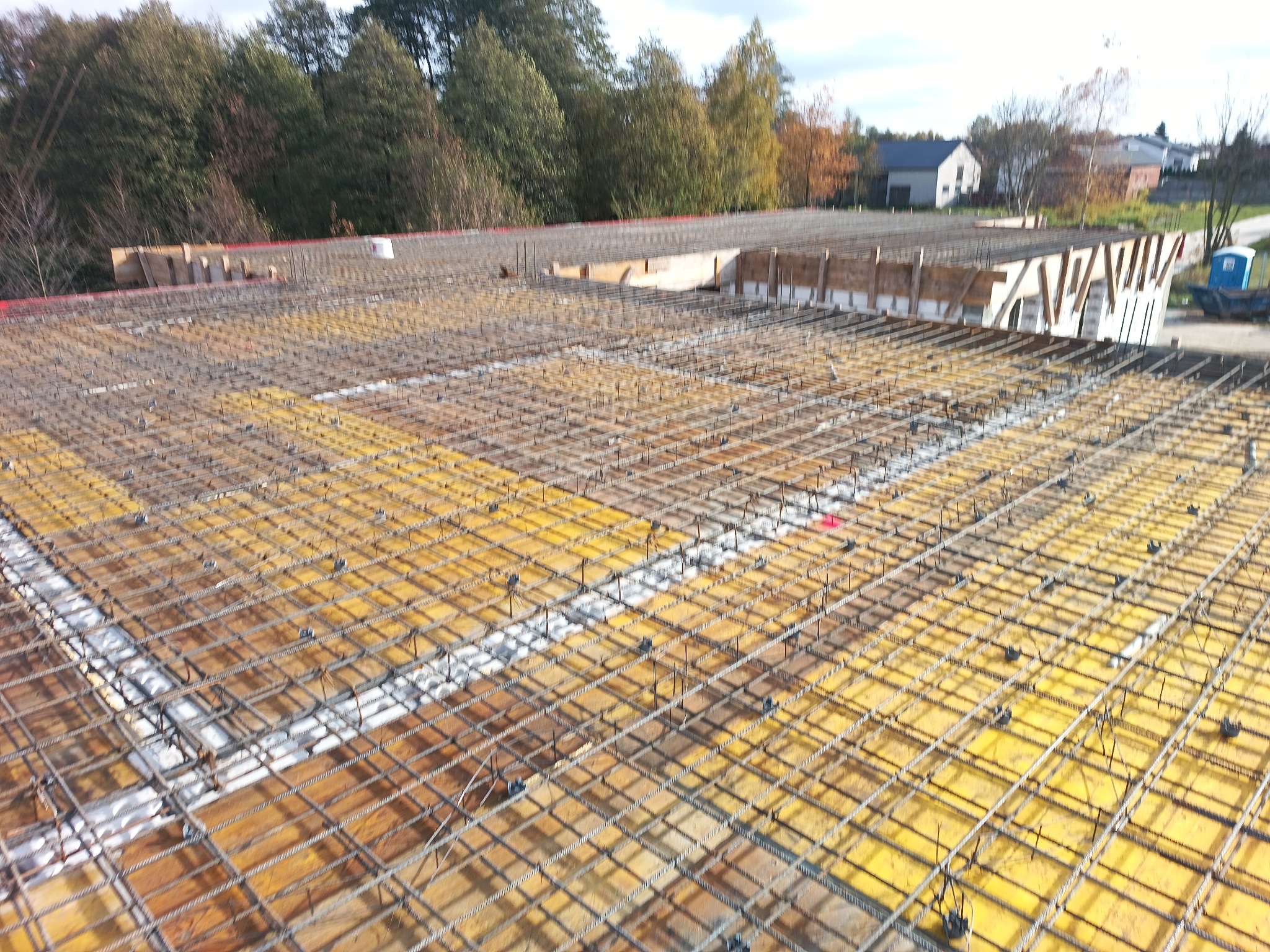 Zbrojenie fundamentu z siatki stalowej na żółtej płycie szalunkowej, widoczne drewniane szalunki krawędziowe i tło z drzewami oraz budynkami.