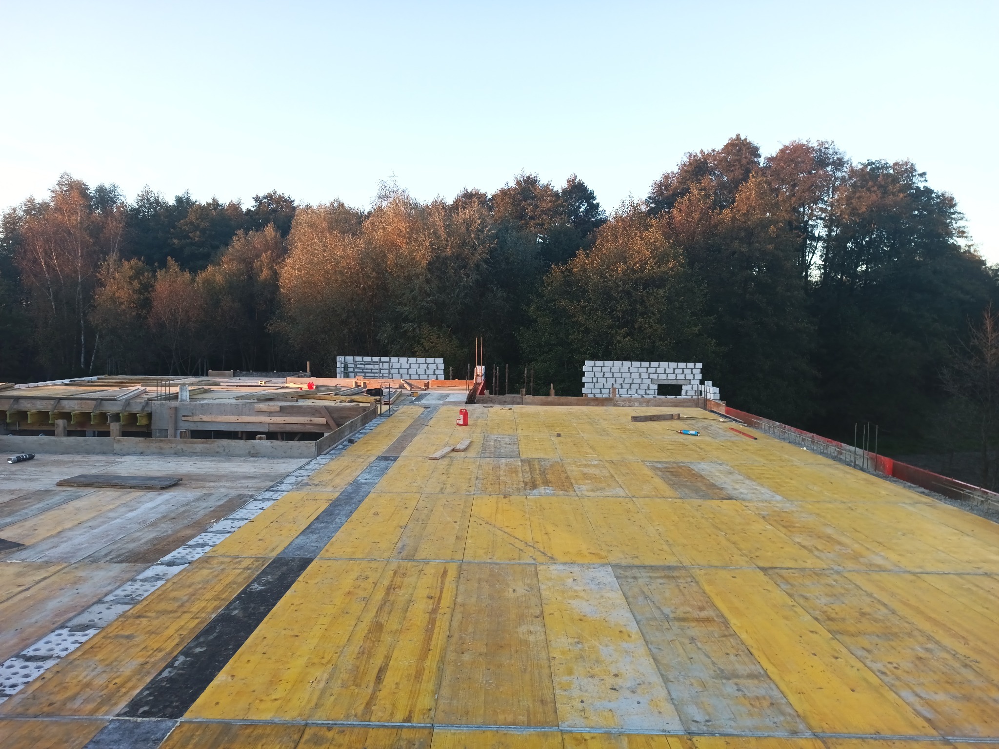 Wczesny etap budowy domu murowanego: widok na fundamenty z żółtym szalunkiem i fragmentem wznoszonych ścian z białych bloczków silikatowych na tle lasu.