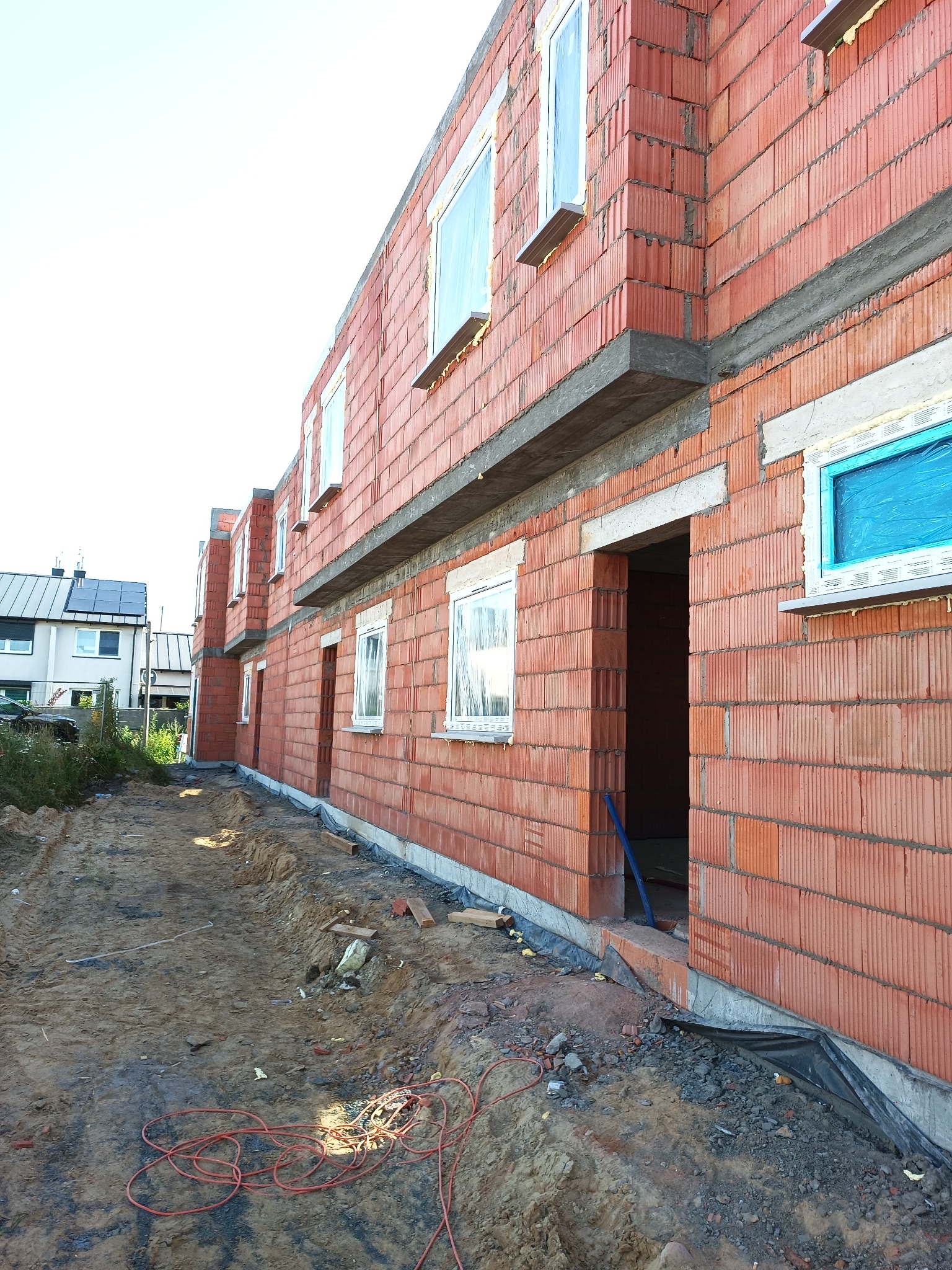 Wznoszenie ścian zewnętrznych z czerwonej cegły ceramicznej porotherm, widoczne okna zabezpieczone folią, stan surowy otwarty, na pierwszym planie teren budowy z przewodami elektrycznymi.