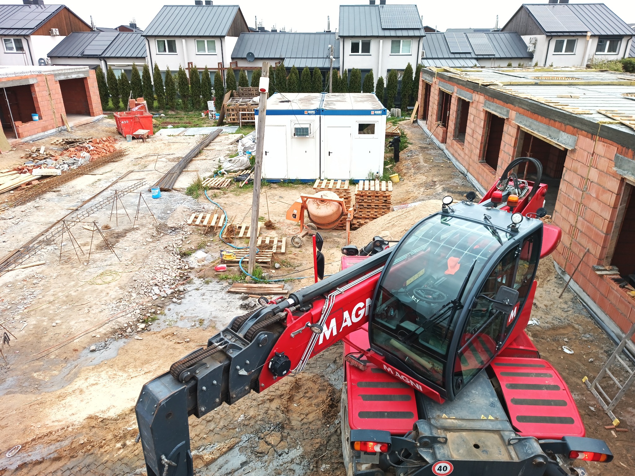 Wysoki kąt widzenia na plac budowy z czerwonym teleskopowym wózkiem widłowym Magni na pierwszym planie, w tle budynki w stanie surowym otwartym, barak budowlany, betonowa gruszka i domy...